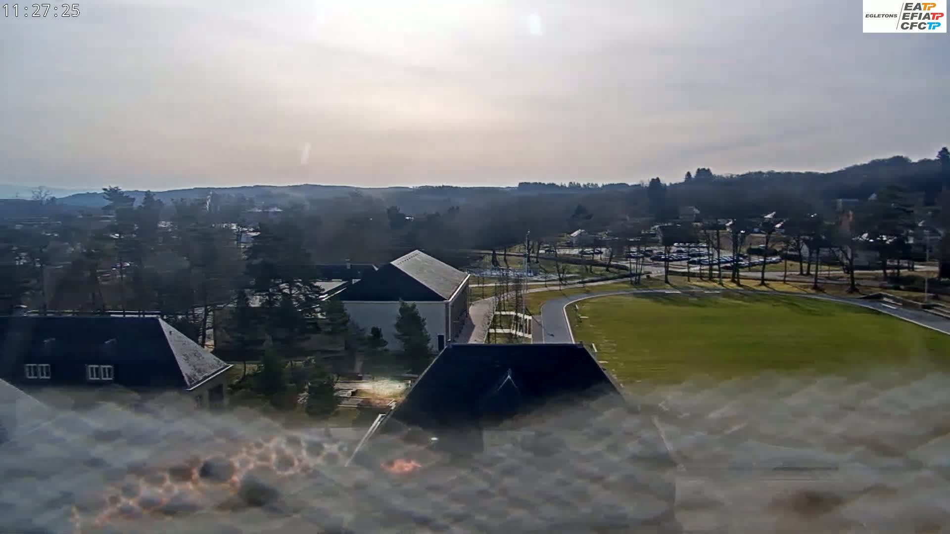 A hazy, sunny day reveals a sprawling landscape featuring buildings, a large grassy field, a parking lot, and a tree-lined hillside in the distance.