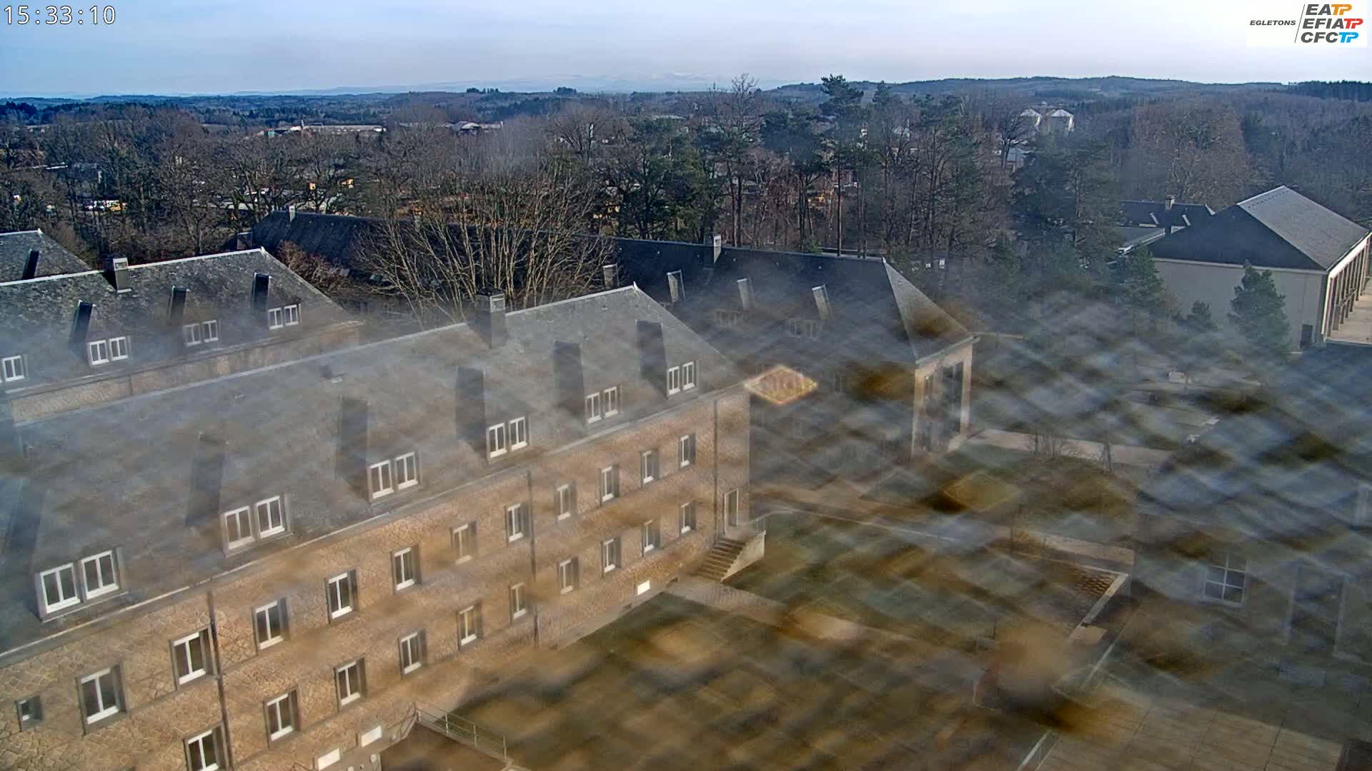 A high-angle, slightly blurry view of several tan stone buildings with dark gray roofs, surrounded by leafless trees on a clear, sunny day.