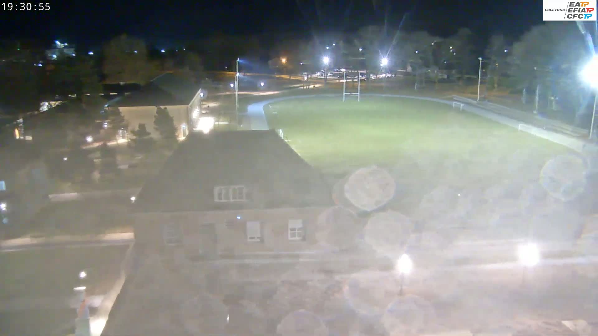 A nighttime, hazy view shows a lit-up athletic field surrounded by buildings and trees.