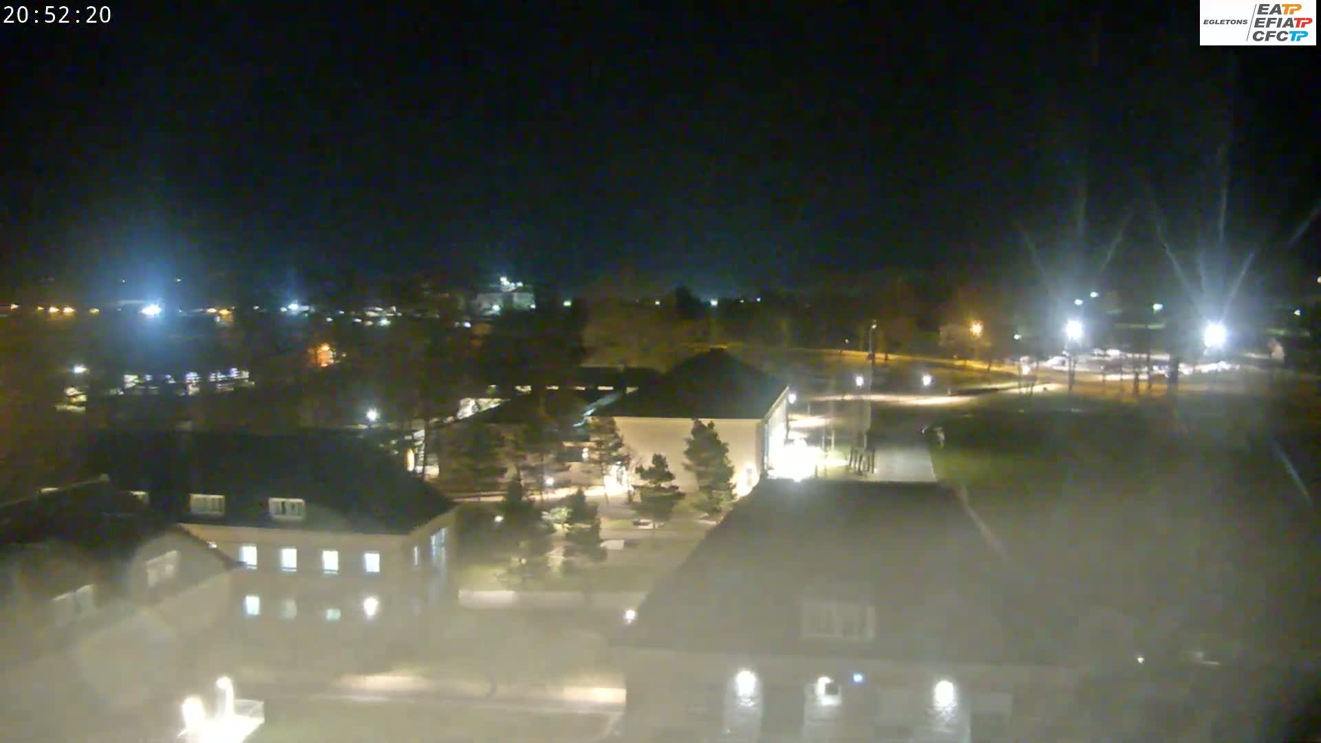 A nighttime aerial view of a campus, showing several buildings with lights on, trees, and a parking lot, under a clear, dark sky.