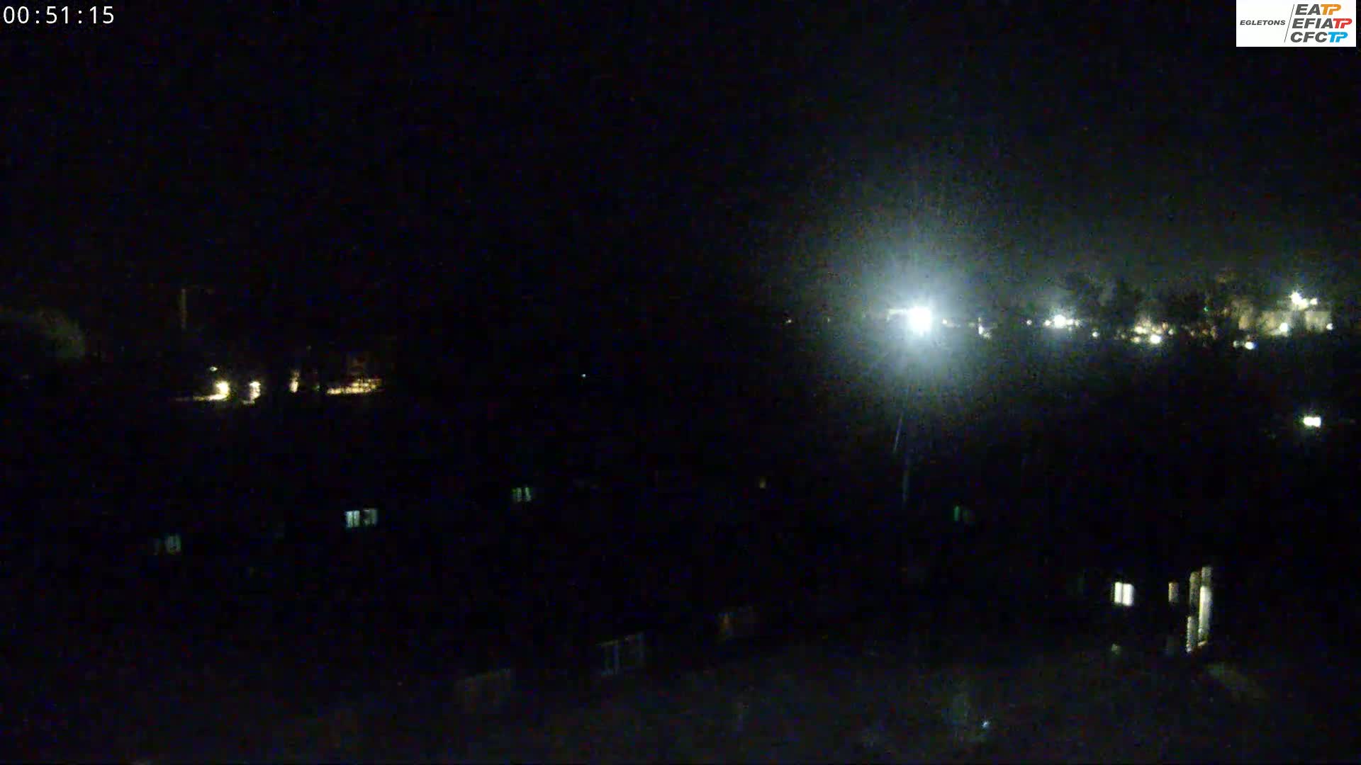 A nighttime, possibly rainy, long shot of a dark residential area with dimly lit windows and distant lights.