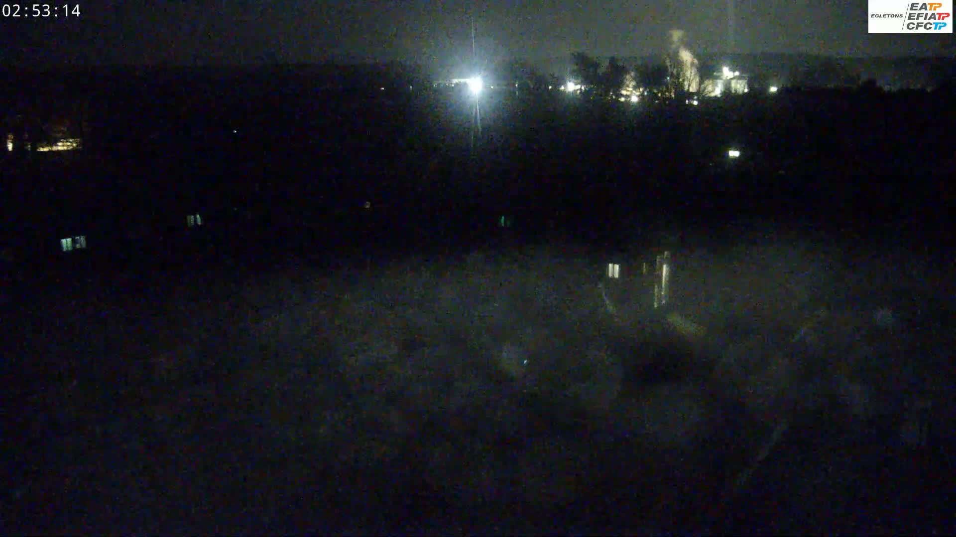 A nighttime, dark view of a landscape with some lights visible in the distance and a few illuminated windows closer to the foreground.