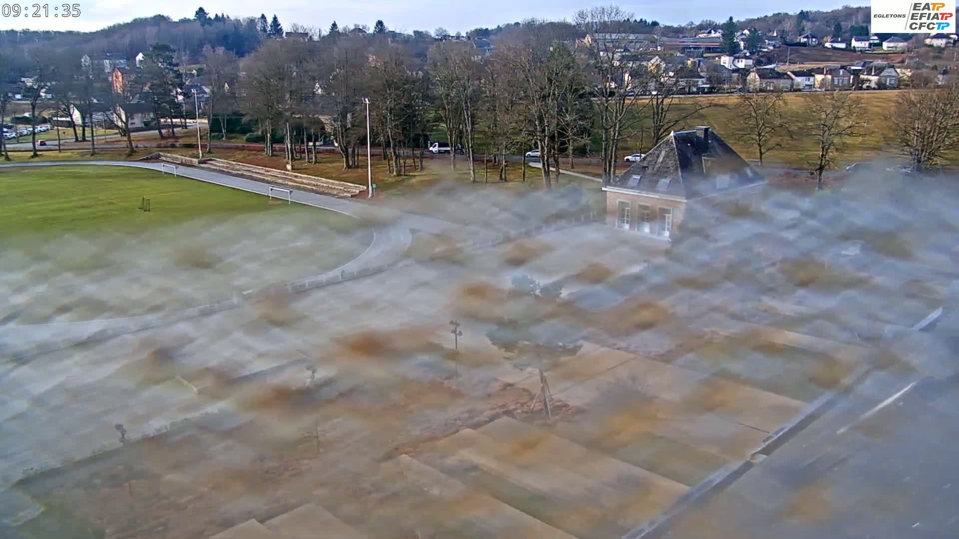 A hazy view of a grassy athletic field, a paved area, a small stone building, and a residential area in the background on a clear day.