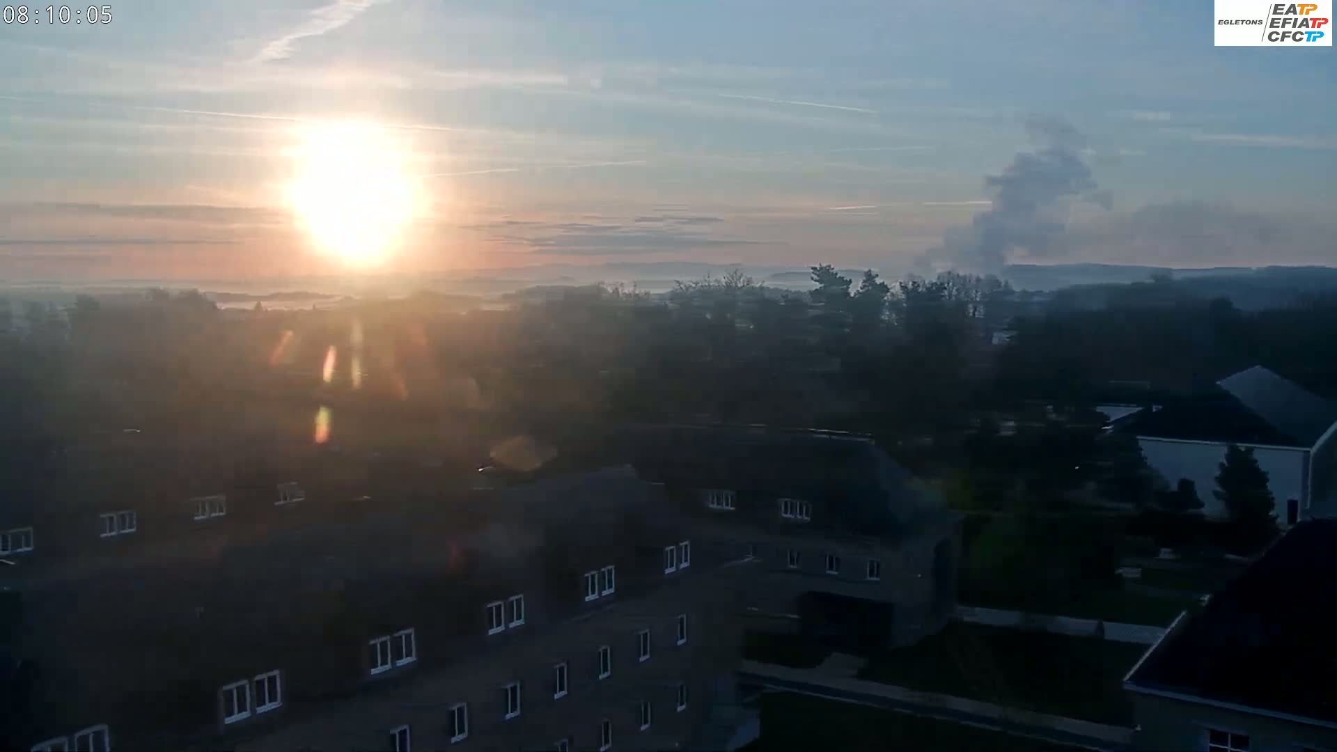 A hazy sunrise illuminates a town of houses and trees shrouded in morning mist, with a plume of smoke visible in the distance.
