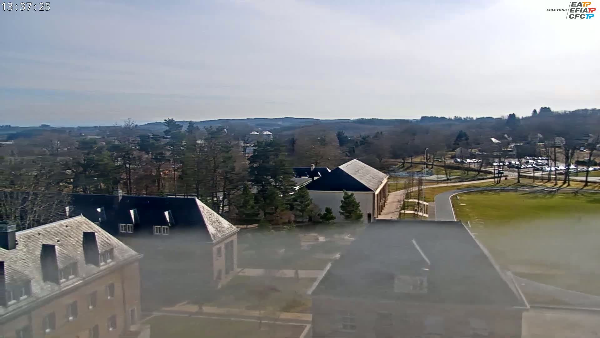 A high-angle, long shot on a hazy day shows several buildings and a large grassy area surrounded by trees and a parking lot.