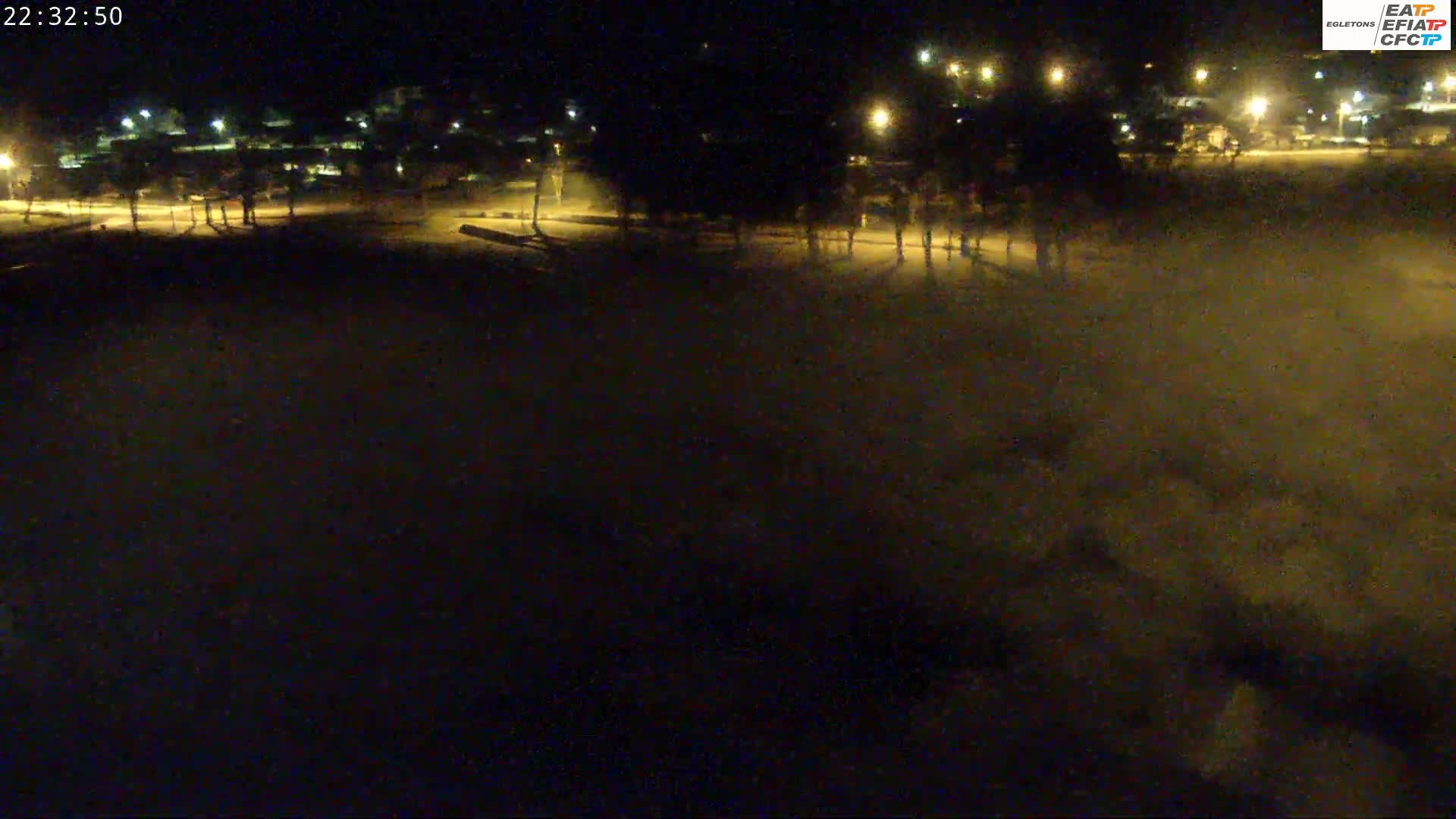 A nighttime, snow-covered field is illuminated by streetlights, with buildings and trees visible in the background.