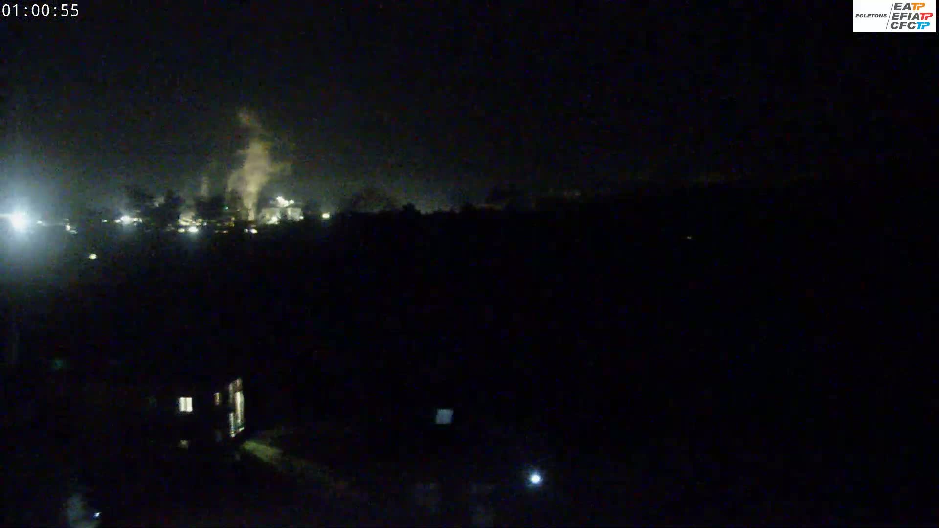 A nighttime, low-light view shows a distant industrial area emitting smoke, with a few lights visible in the foreground.