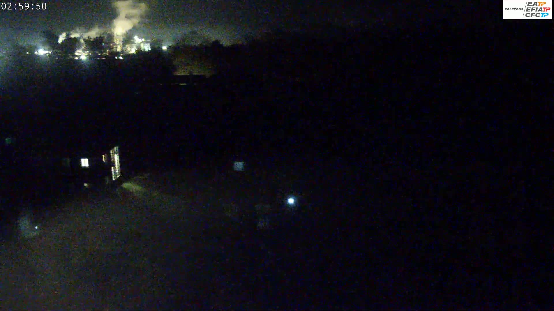A nighttime view shows a dimly lit industrial area with smoke plumes, a dark landscape, and a few small lights in the foreground.