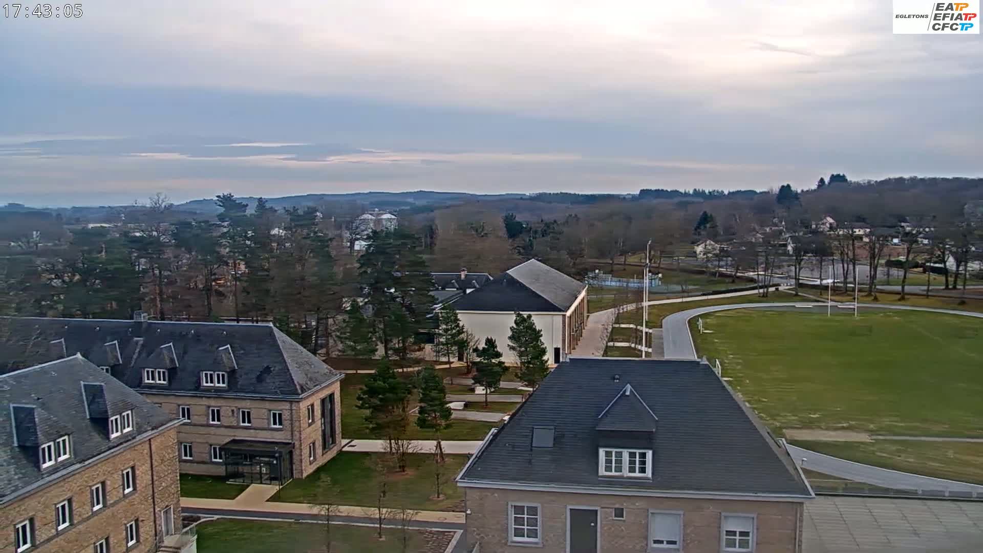 An overcast day reveals a campus with several buildings, a running track, and a grassy field, all surrounded by winter trees and distant hills.