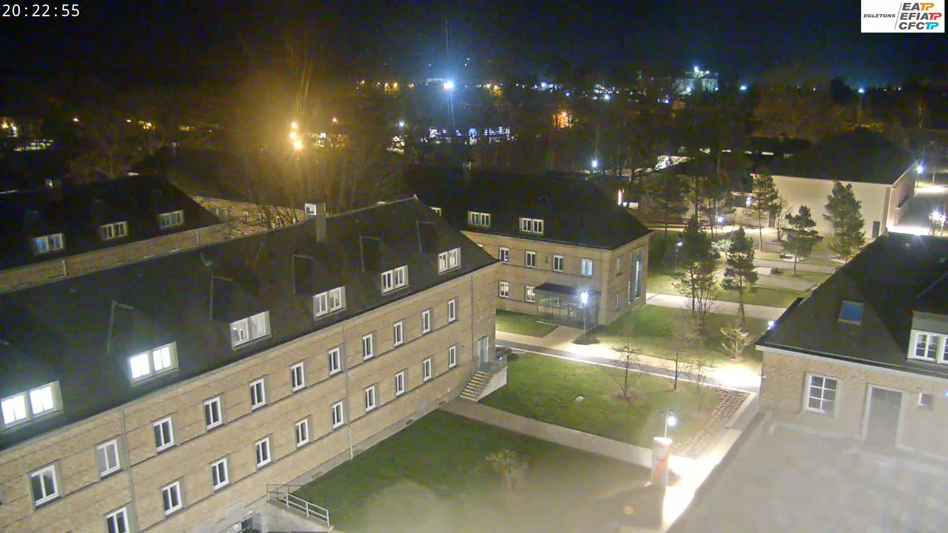 A nighttime aerial view of a complex of low-rise buildings with lit windows, surrounded by trees and paved walkways.