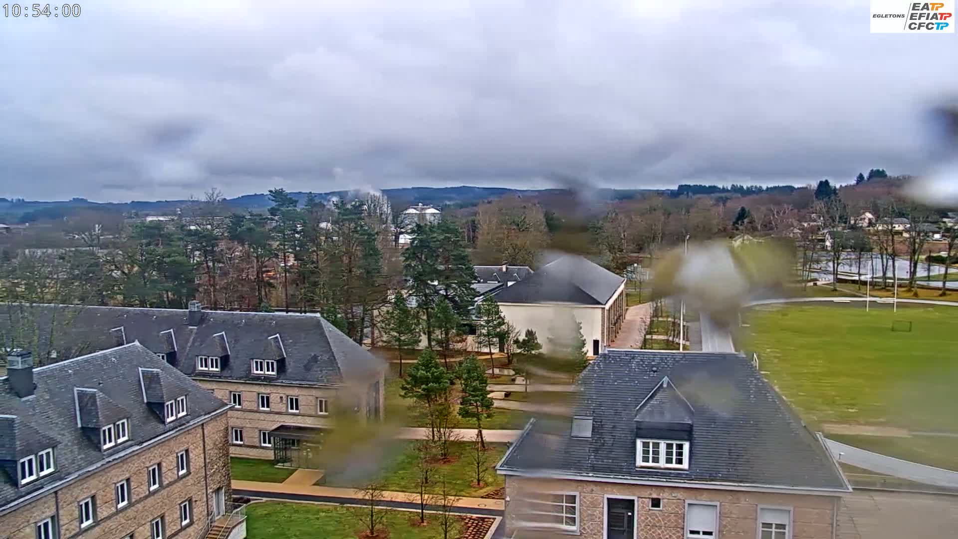 A rain-streaked view of several stone buildings, a grassy field, and trees under an overcast sky.