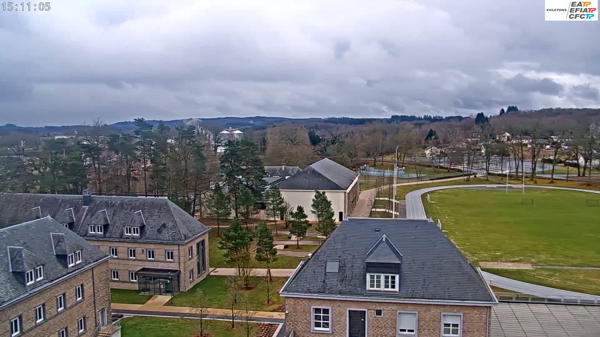 An aerial view shows a campus with several stone buildings, a running track, and a large grassy field under an overcast sky.