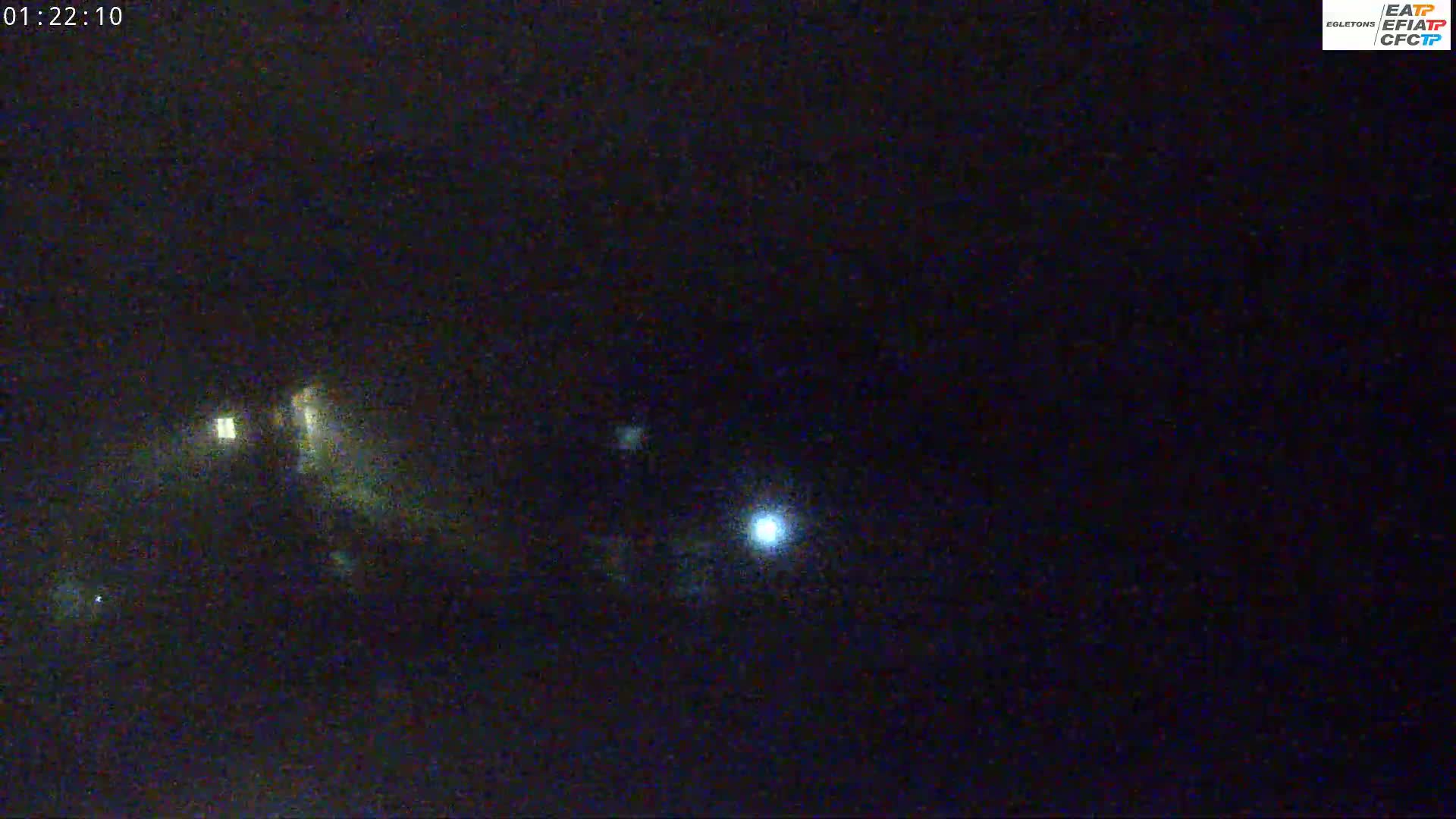 A nighttime, low-light image shows a dark scene with a few distant lights, possibly vehicle headlights, and a brighter light source in the lower center that could be a streetlight or a similar object.