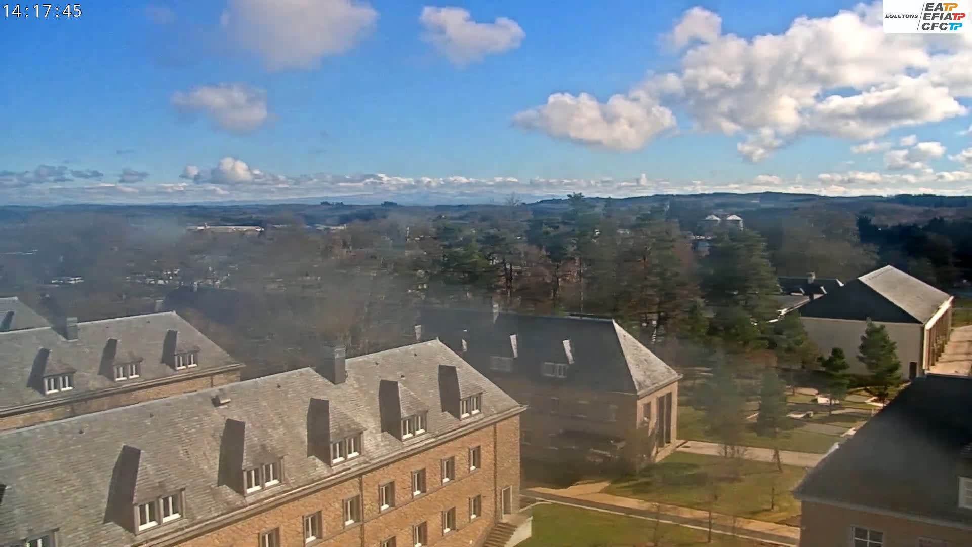A hazy, partly sunny day reveals a campus scene of several stone buildings with grey slate roofs, surrounded by trees, and distant rolling hills under a blue sky with fluffy white clouds.