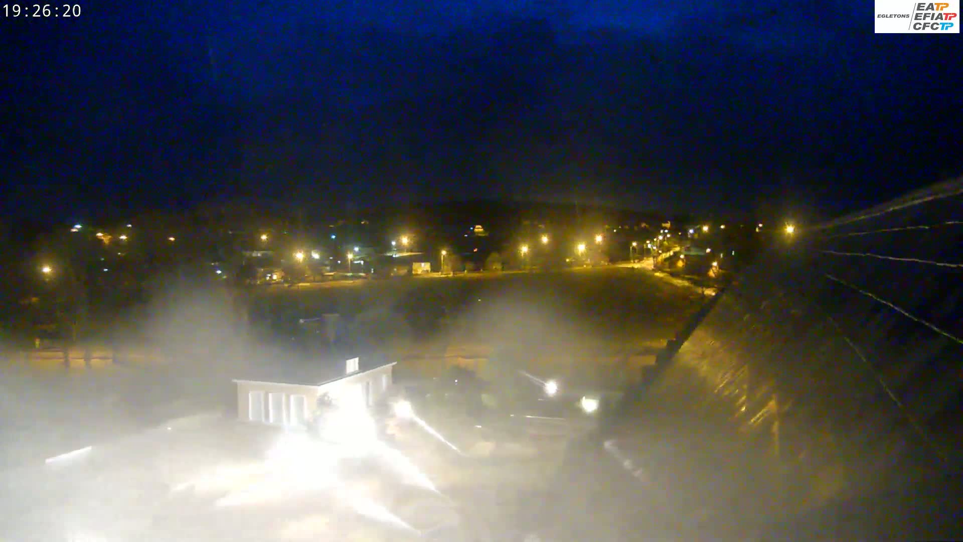 A nighttime view of a large structure, possibly a dam, with water cascading over it, creating mist, and a smaller building visible in the foreground, under a dark sky.