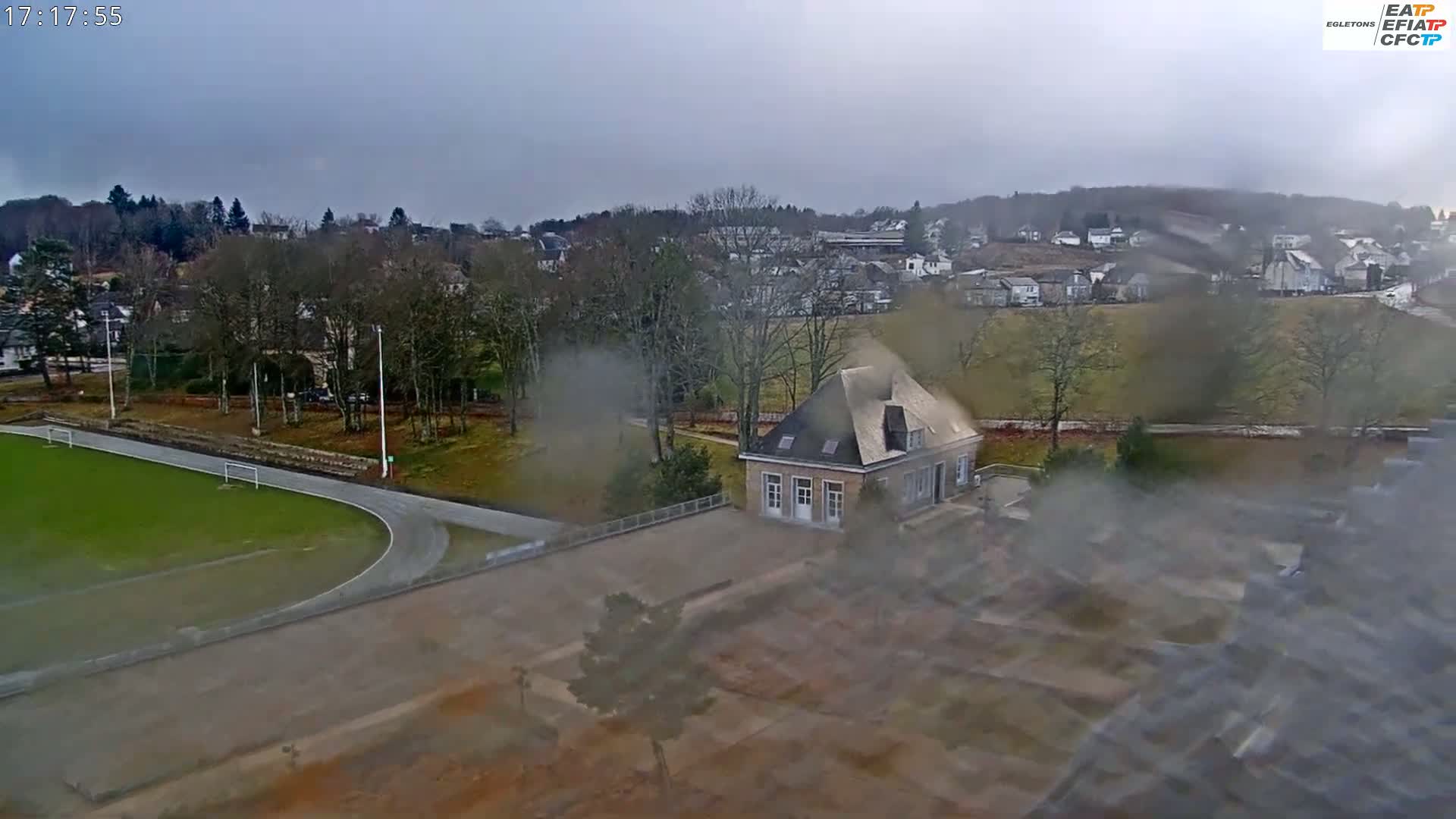 A high-angle, long shot on an overcast day shows a small stone building near a running track and soccer field, with a residential area and trees in the background.