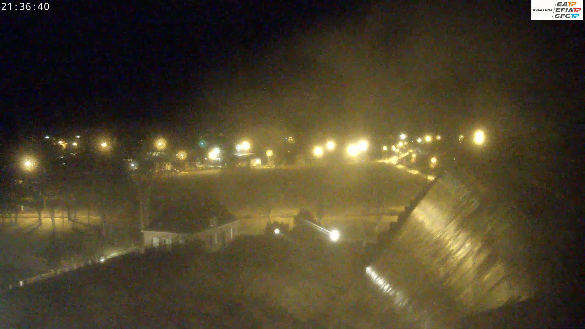 A nighttime view of a dam with water flowing over it, illuminated by streetlights across a dark, possibly foggy or hazy, landscape.
