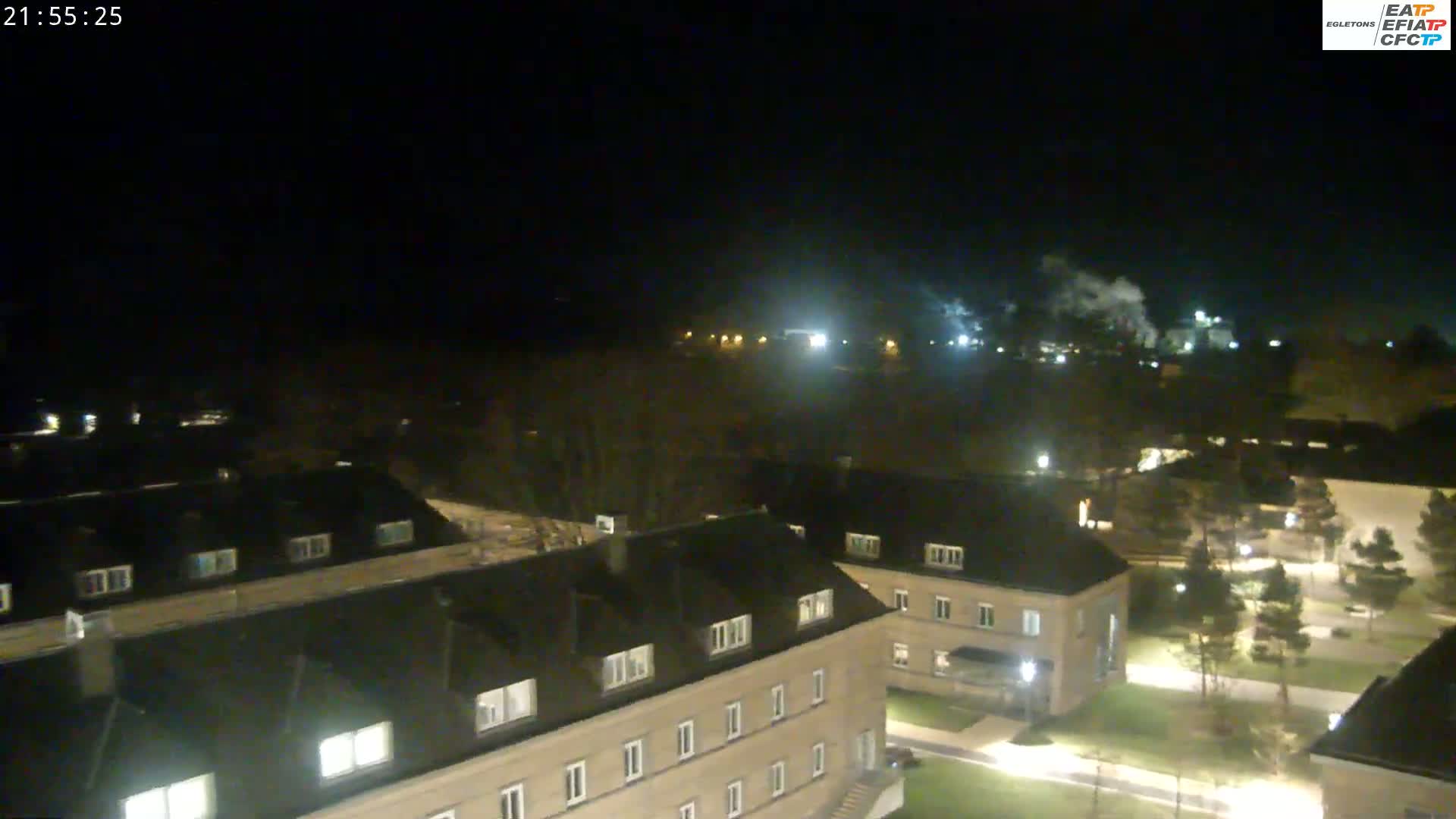 A nighttime aerial view of a residential area with several buildings illuminated, some smoke visible in the distance under a dark sky.
