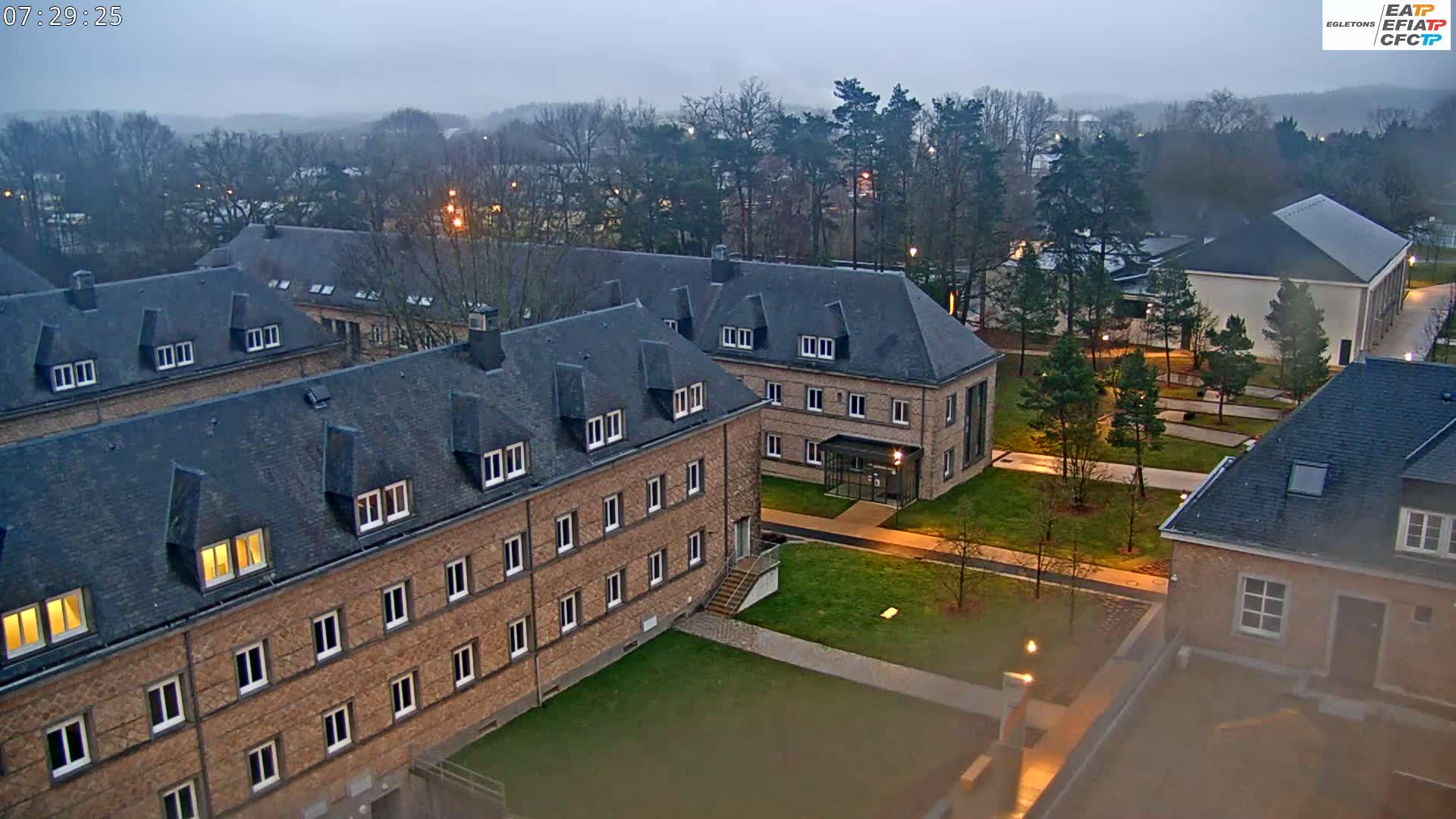 An aerial view of a campus under an overcast sky shows several brick buildings with slate roofs and manicured lawns and walkways between them.
