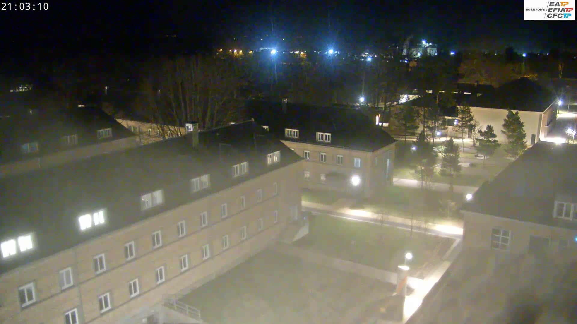 An aerial nighttime view of a complex of buildings with lit windows, surrounded by trees and paved pathways, under a dark sky.