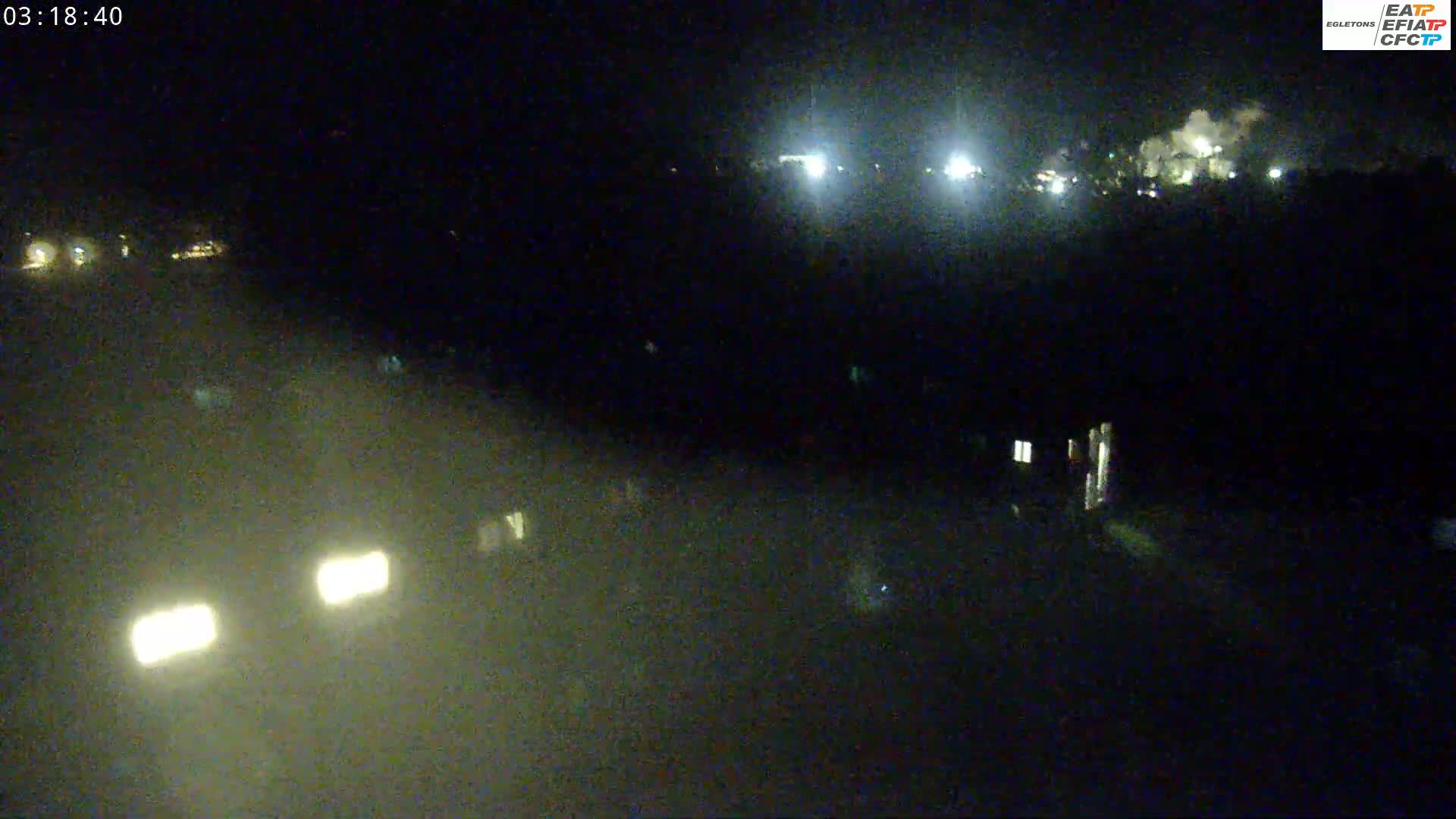 At night, vehicle headlights illuminate a dark, hazy scene with distant lights and faint smoke or dust in the air.
