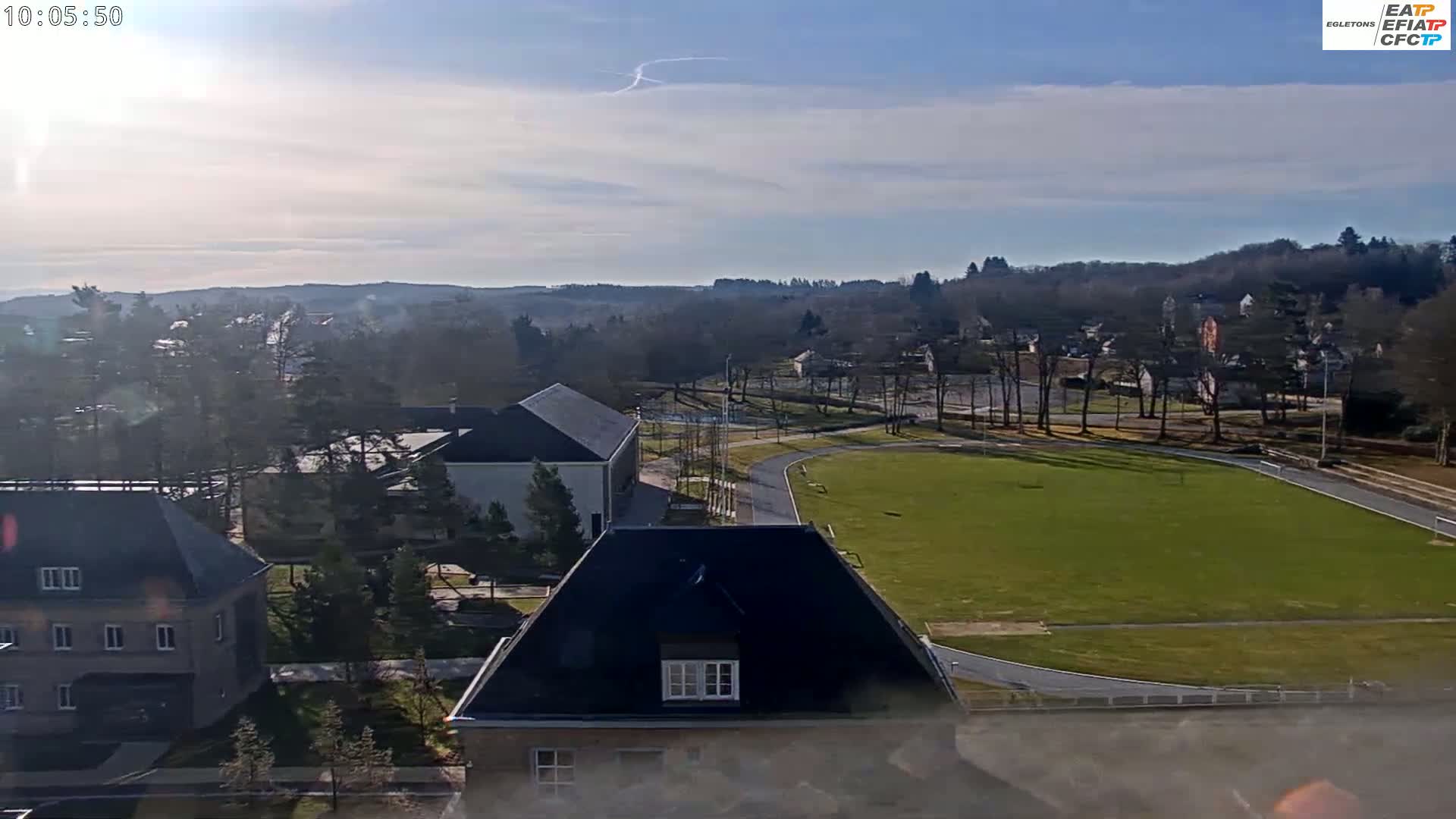 A high-angle, long shot on a sunny day shows a grassy athletic field surrounded by buildings and trees, with hills visible in the distance.