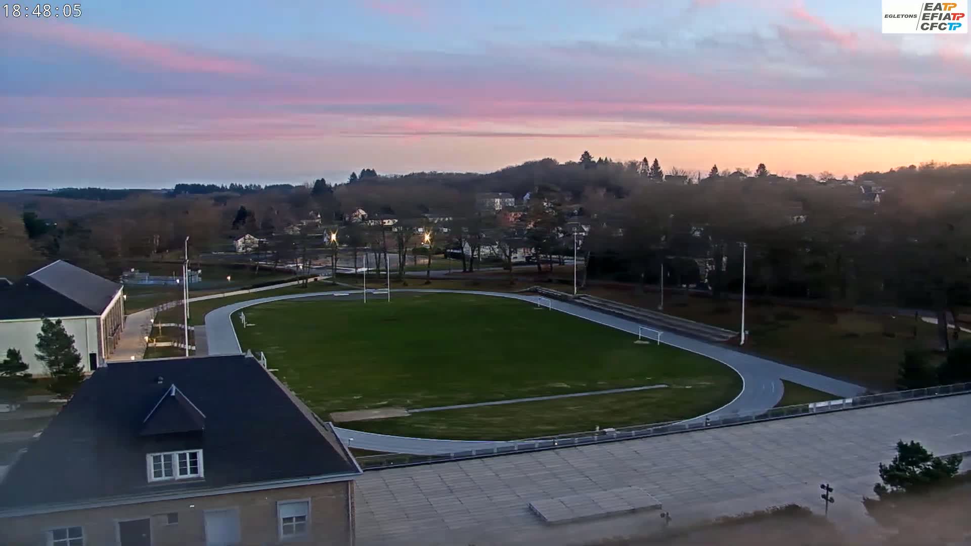 A high-angle view shows a grassy athletic field with a running track, surrounded by buildings and trees under a twilight sky with pinkish hues.