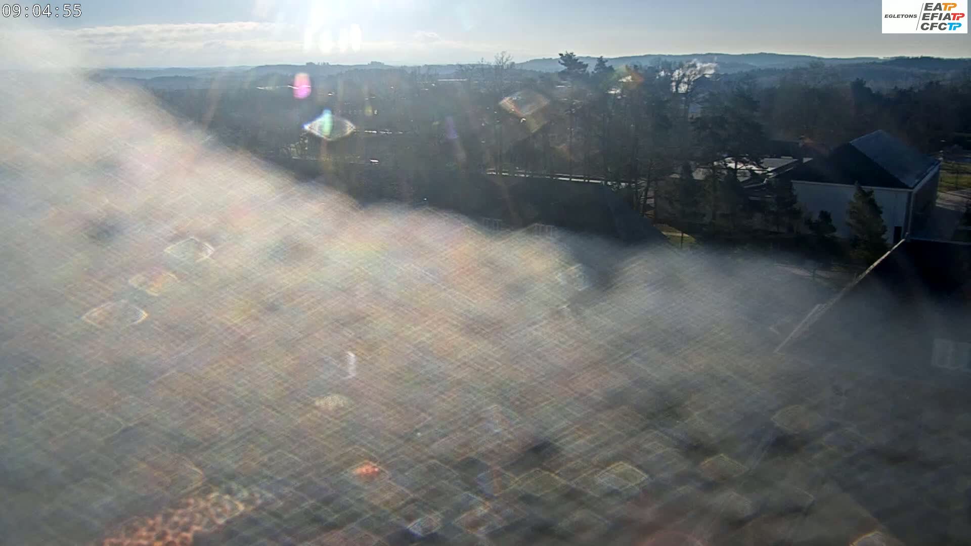 A hazy, sun-dappled view of a suburban area with buildings and trees on a partly cloudy day.