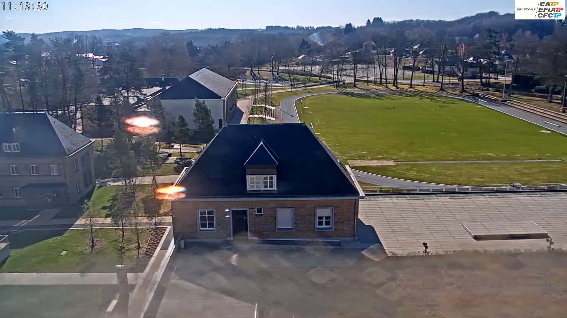 A sunny day reveals a brick house and a grassy athletic field surrounded by buildings and trees.