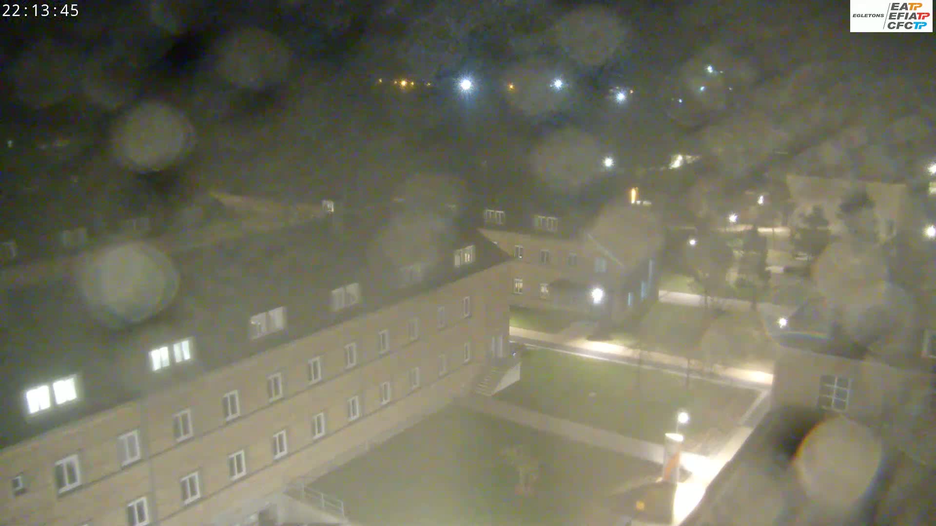 A nighttime, rain-blurred view shows a multi-story building complex with grassy areas and walkways between the structures.