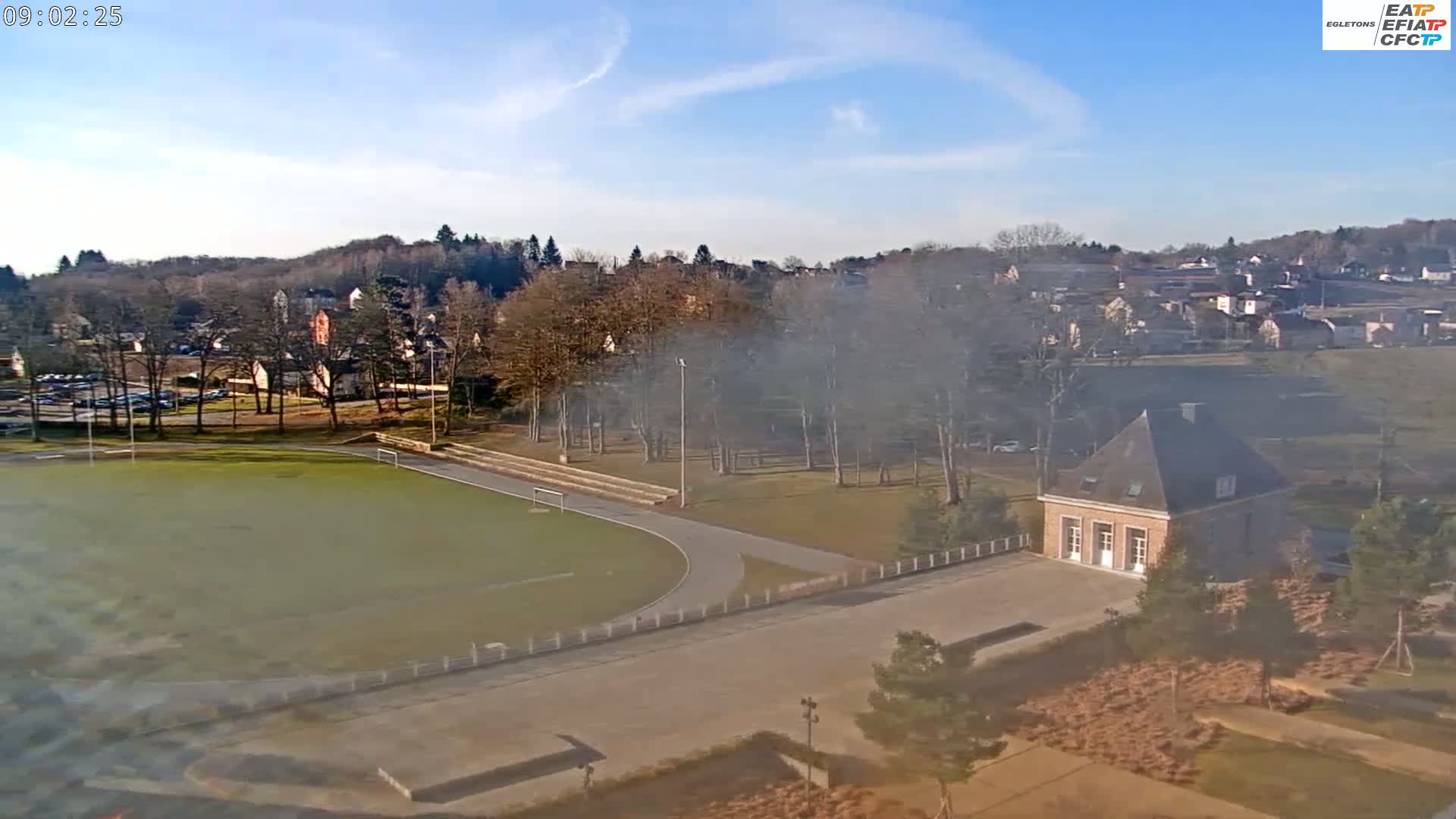 An elevated view of a sports field, a small building, and a residential area on a hazy, sunny day.