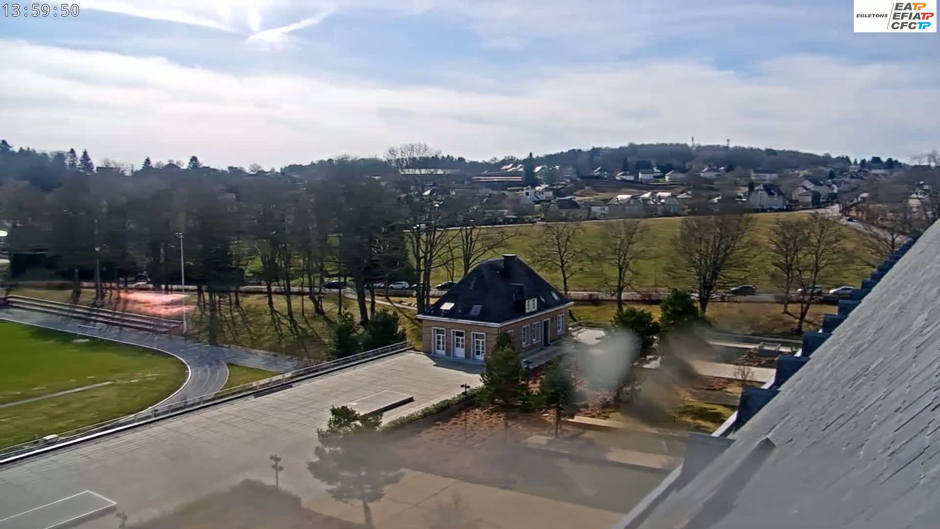 A small, light-brown building sits on a paved area next to a running track and soccer field, overlooking a town nestled amongst bare trees under a mostly sunny sky.