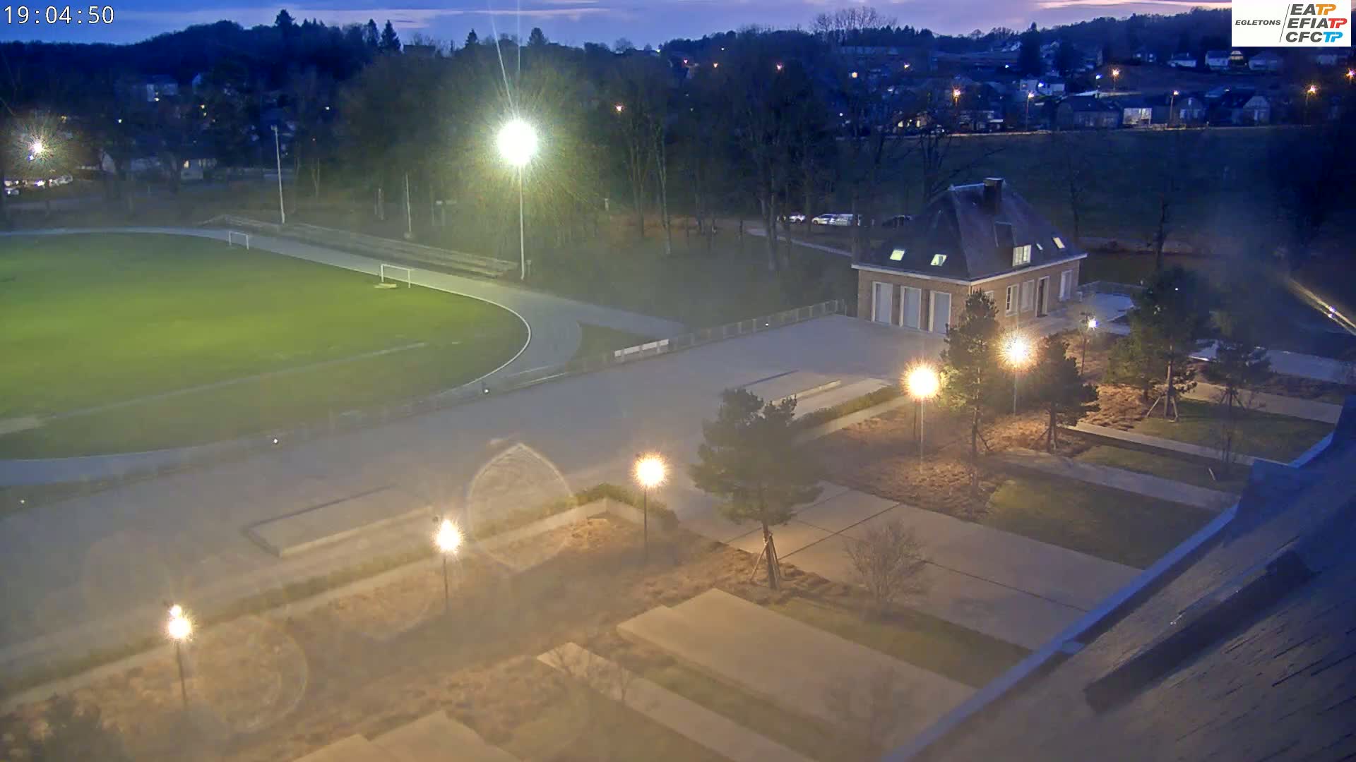 A nighttime aerial view of a sports field with a running track, a building, and landscaped grounds illuminated by artificial lights under a twilight sky.