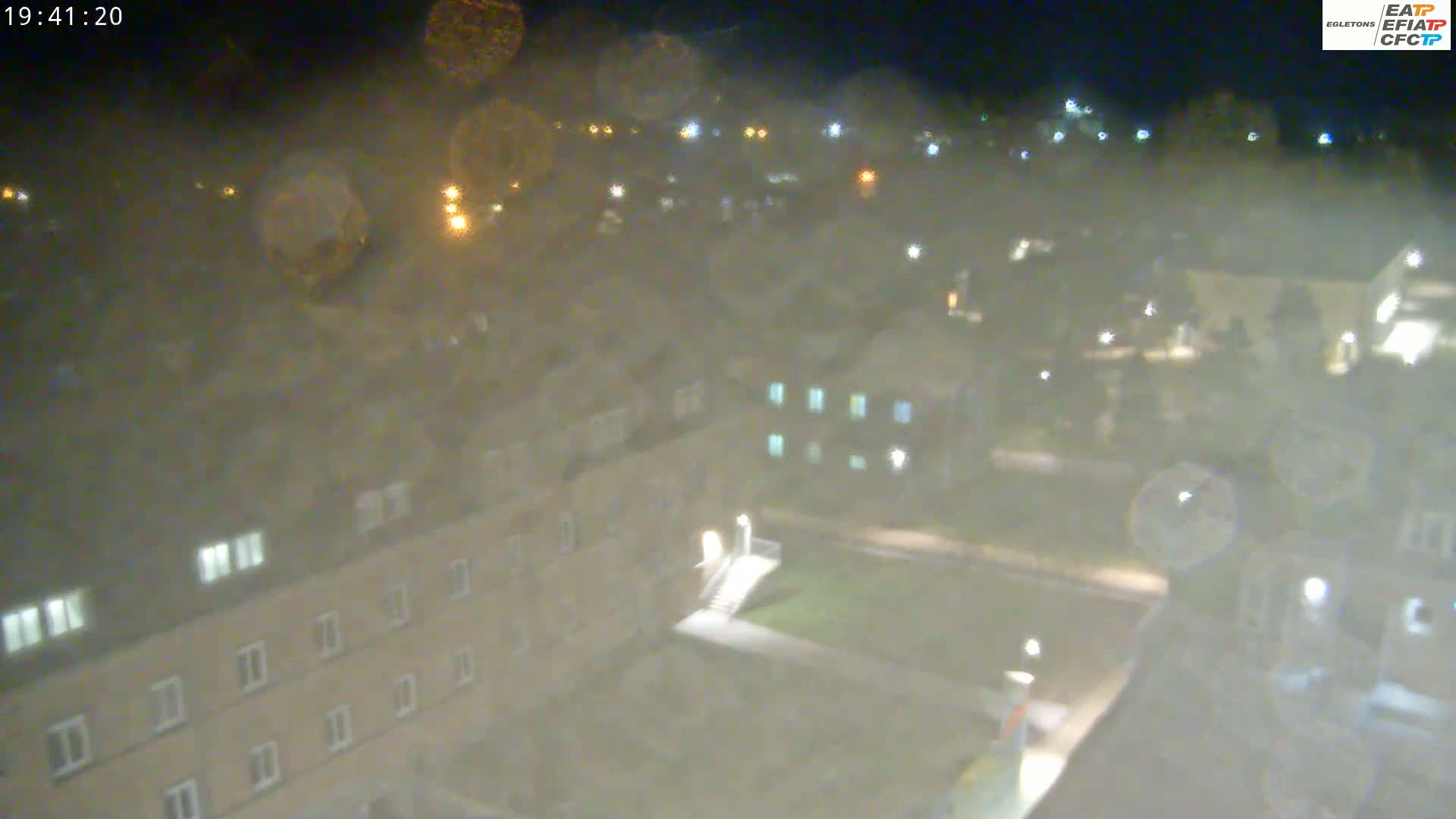 A nighttime, high-angle view of a campus with several buildings and a grassy area, obscured by light rain or mist.