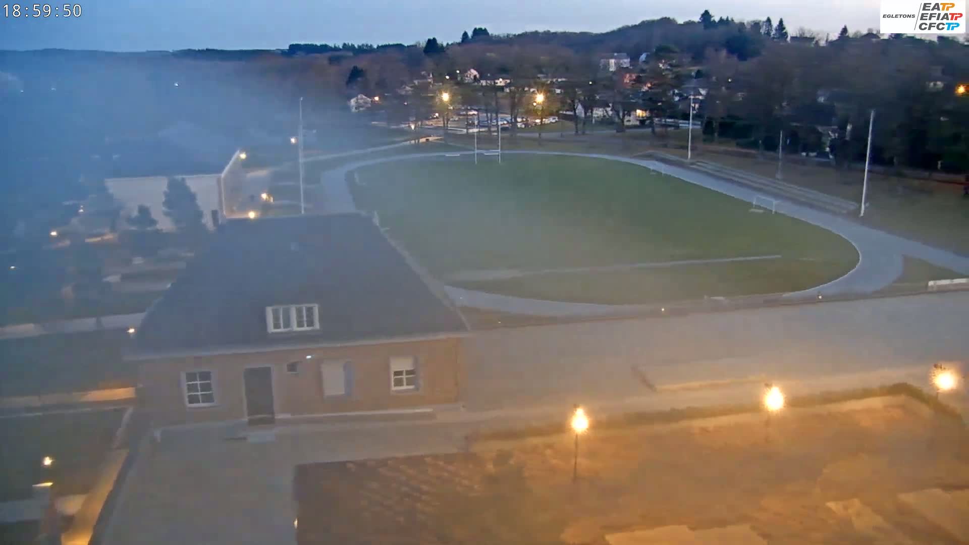 A hazy evening view shows a running track and soccer field, next to a building, with lights illuminating the scene.