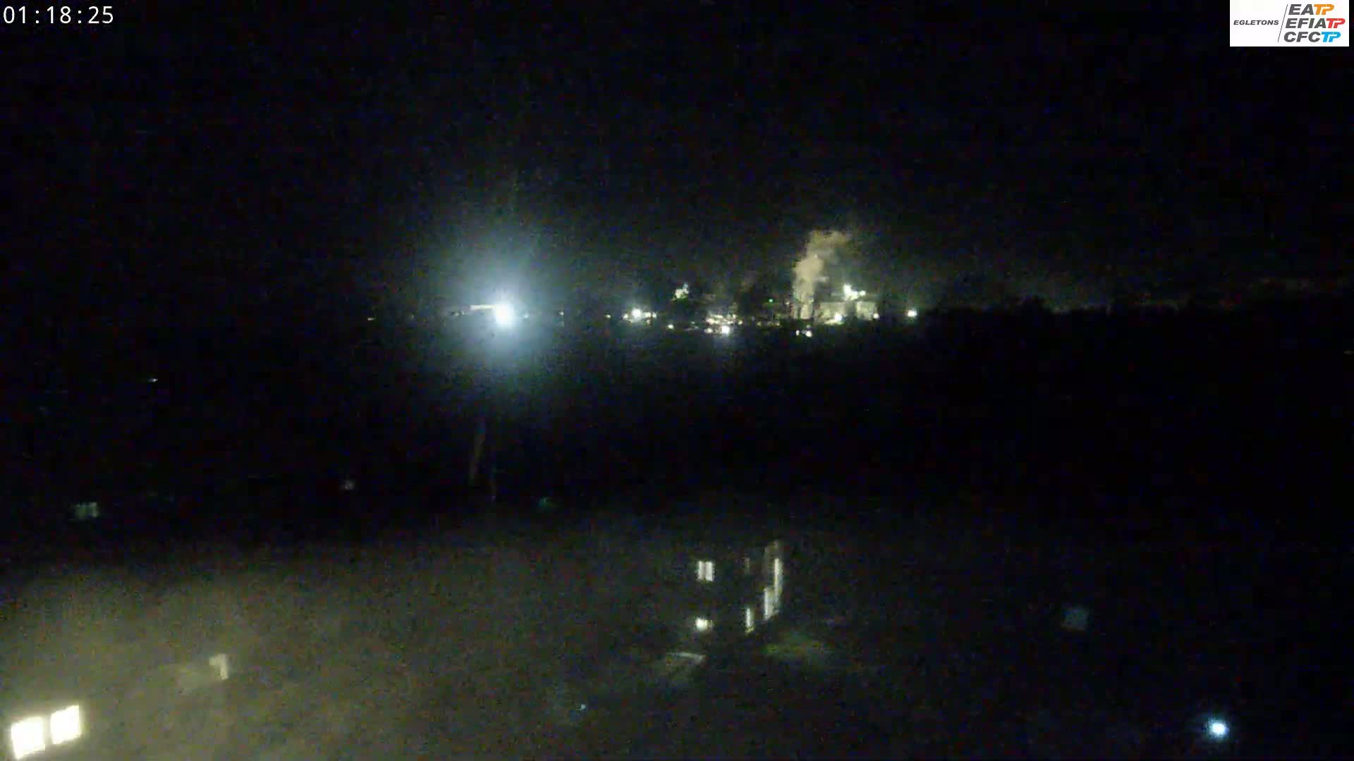 A nighttime long shot shows an industrial area with lights and smoke emitting from a structure, under a dark, clear sky.