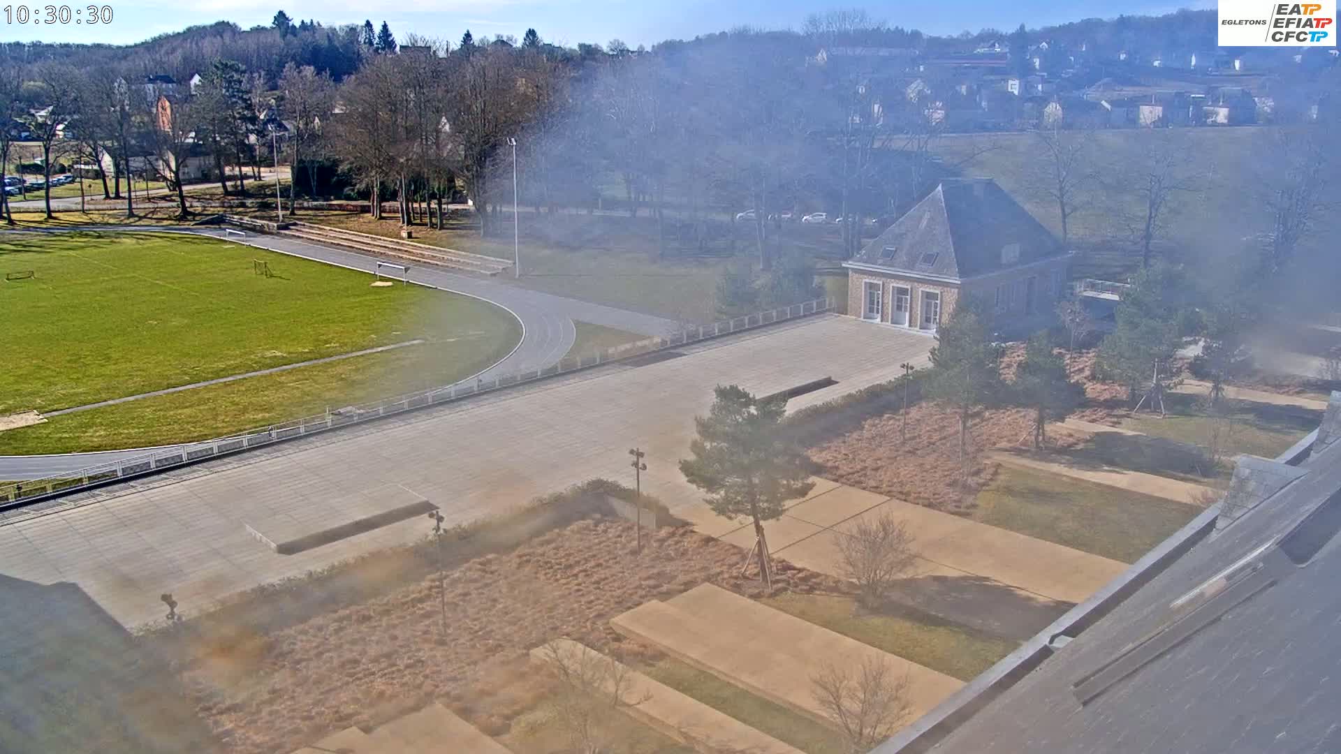An aerial view shows a sports complex with a running track and grassy field, adjacent to a courtyard with landscaped areas and a stone building, all under a hazy sunny sky.