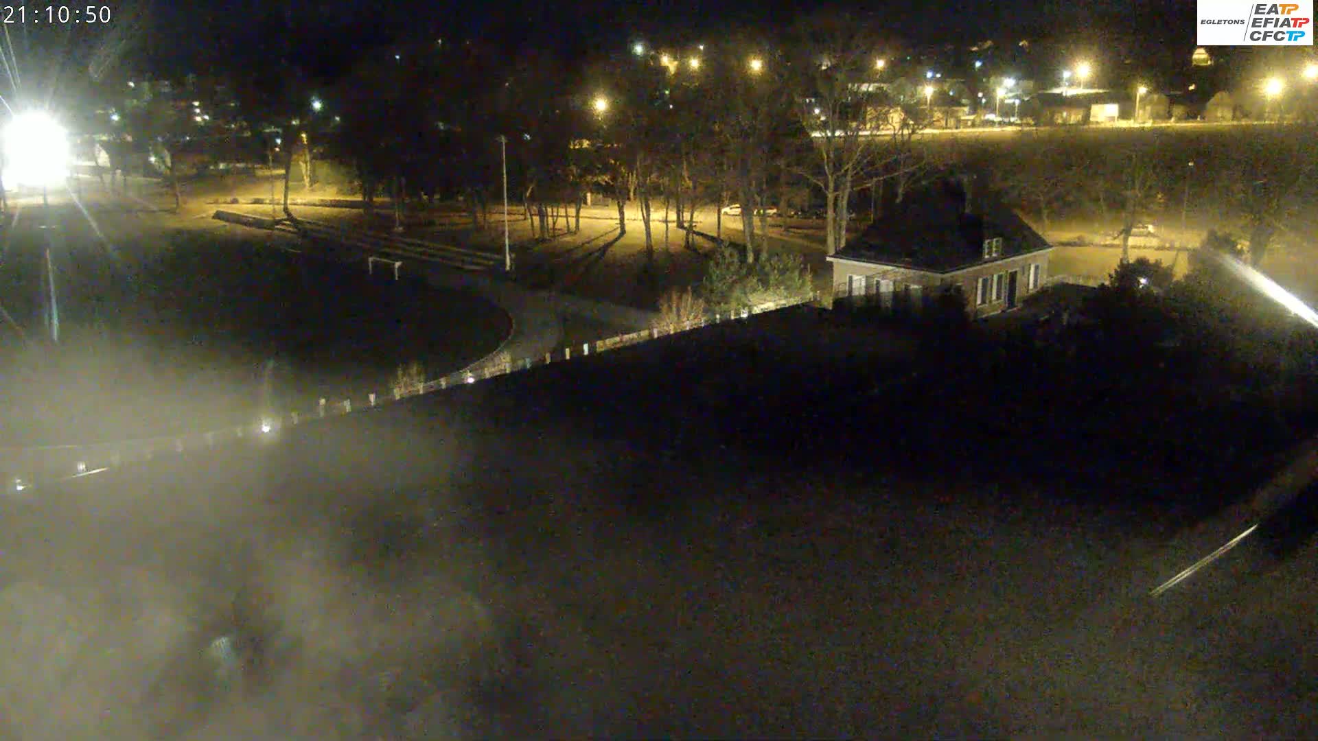 A nighttime aerial view shows a park with a small building, trees, and a paved area, under clear, dark skies.