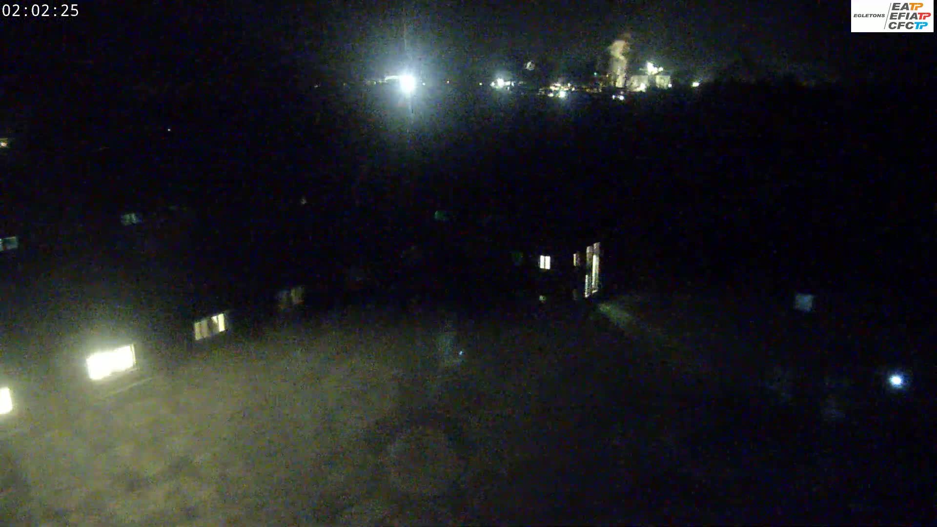 A nighttime, high-angle view shows a dark landscape with dimly lit buildings and a brighter area in the distance, possibly industrial, under a clear, dark sky.