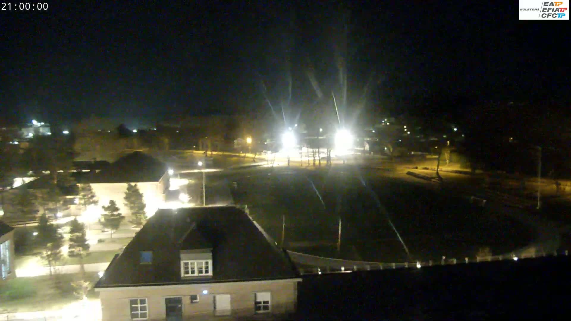 An aerial nighttime view shows a dark, empty field surrounded by buildings and trees, illuminated by streetlights and car headlights.