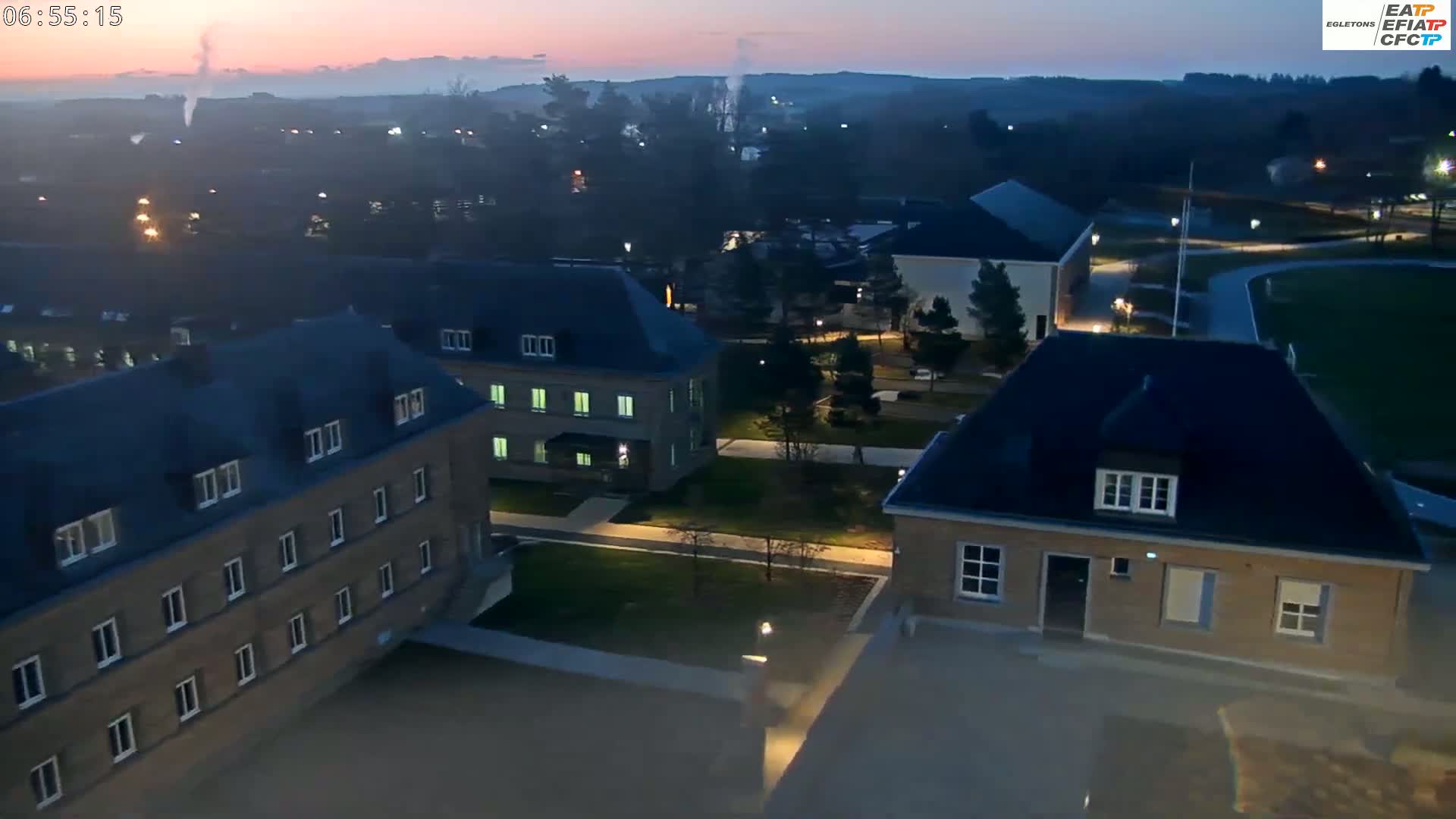 An aerial view at dawn shows several low-lying buildings with dark roofs and light-colored walls in a campus-like setting, with trees and grassy areas visible.