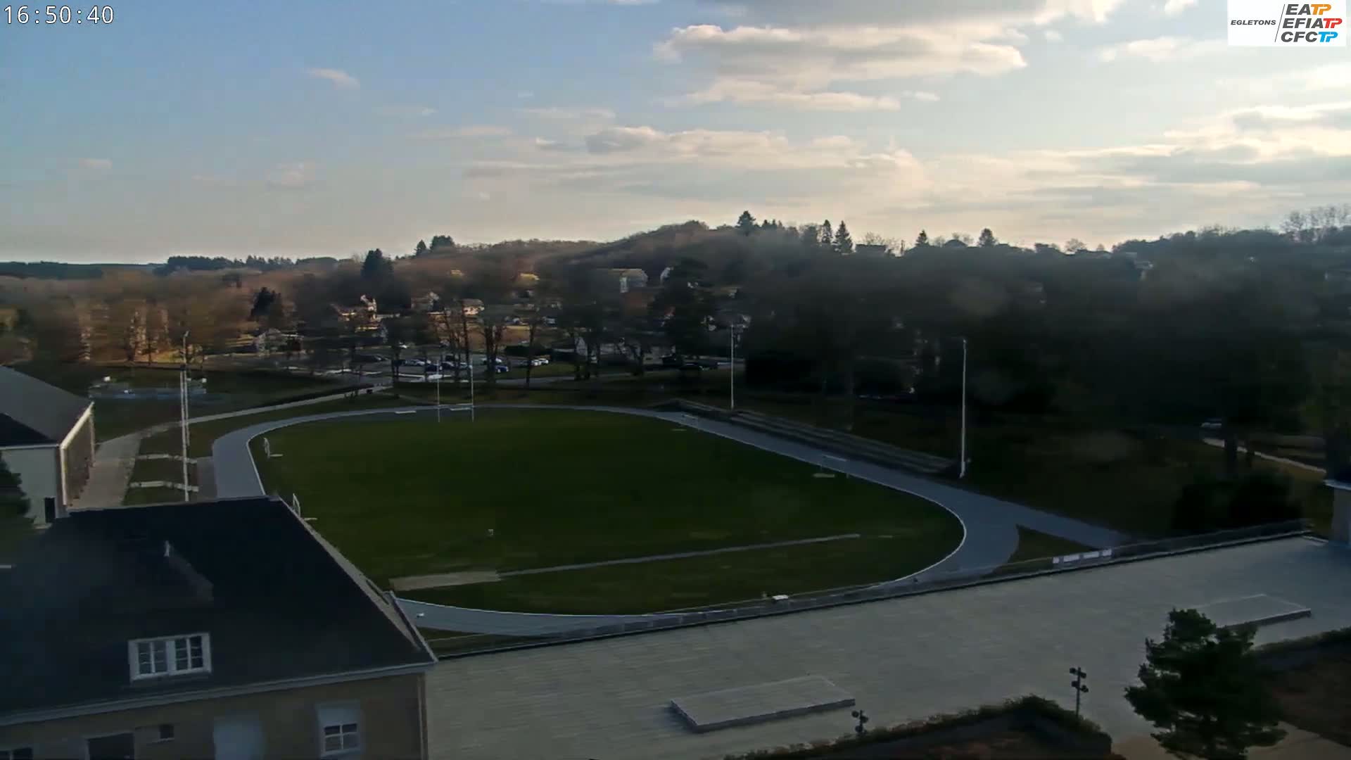An outdoor view of an oval-shaped athletic field with a running track, surrounded by buildings and trees under a mostly clear sky.