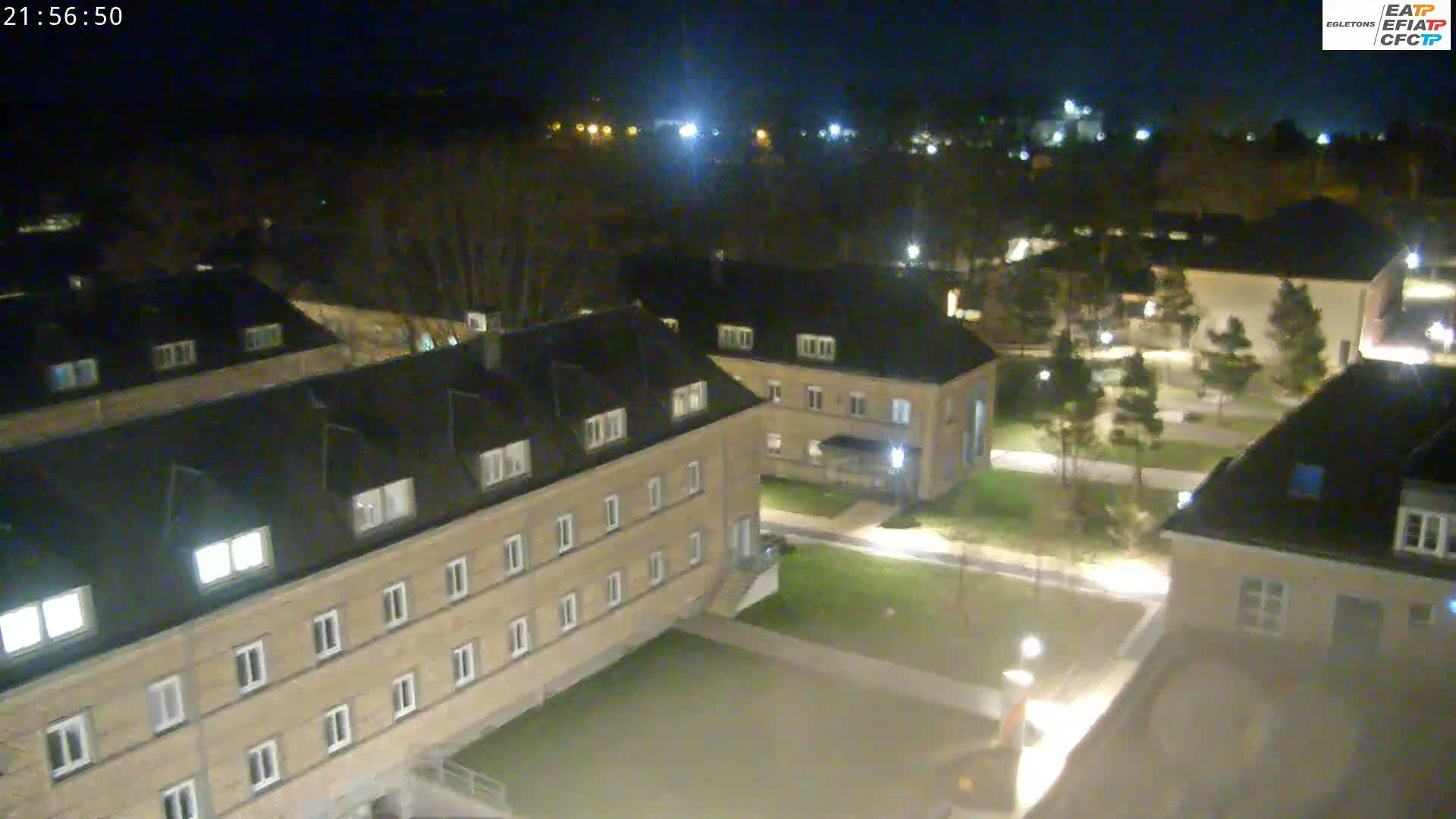 An aerial nighttime view shows several low-rise buildings with illuminated windows, situated on a paved courtyard with some grassy areas and trees.