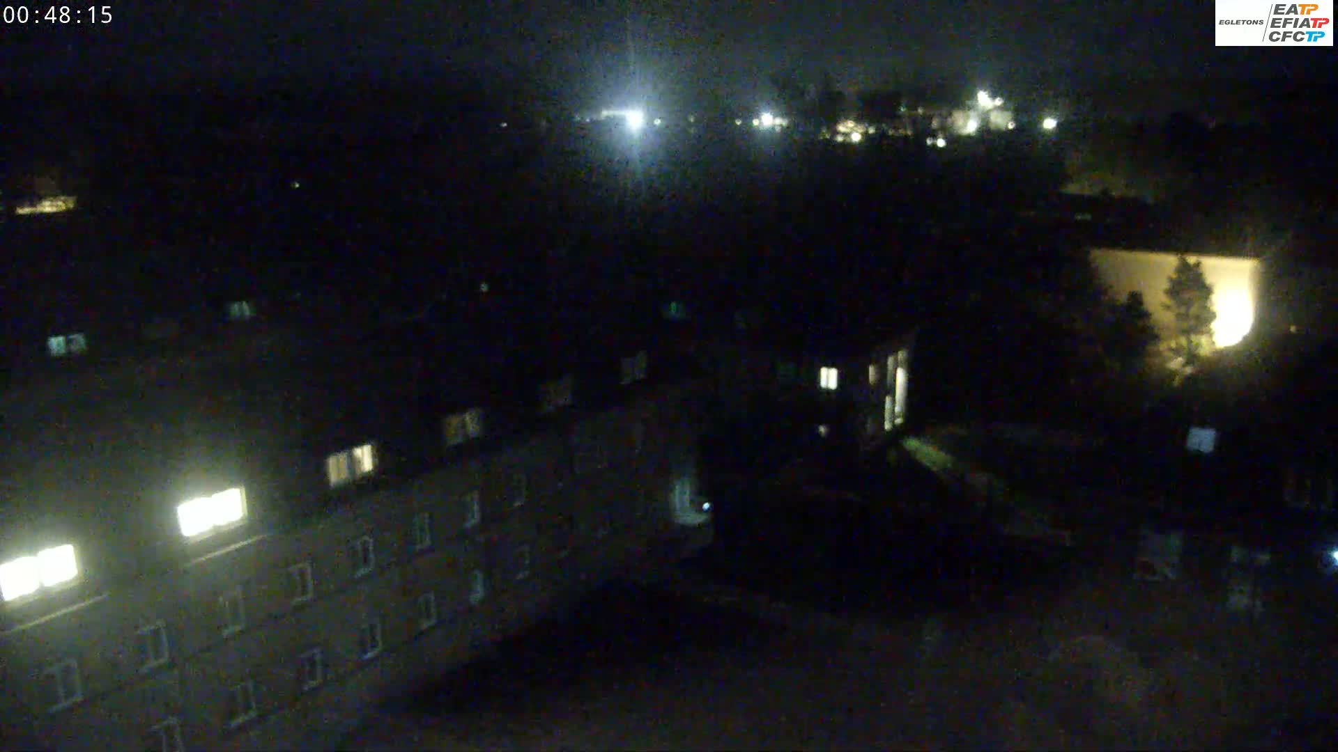 A nighttime, high-angle view shows a multi-story building with some illuminated windows, surrounded by dark, sparsely lit grounds and other buildings in the distance.
