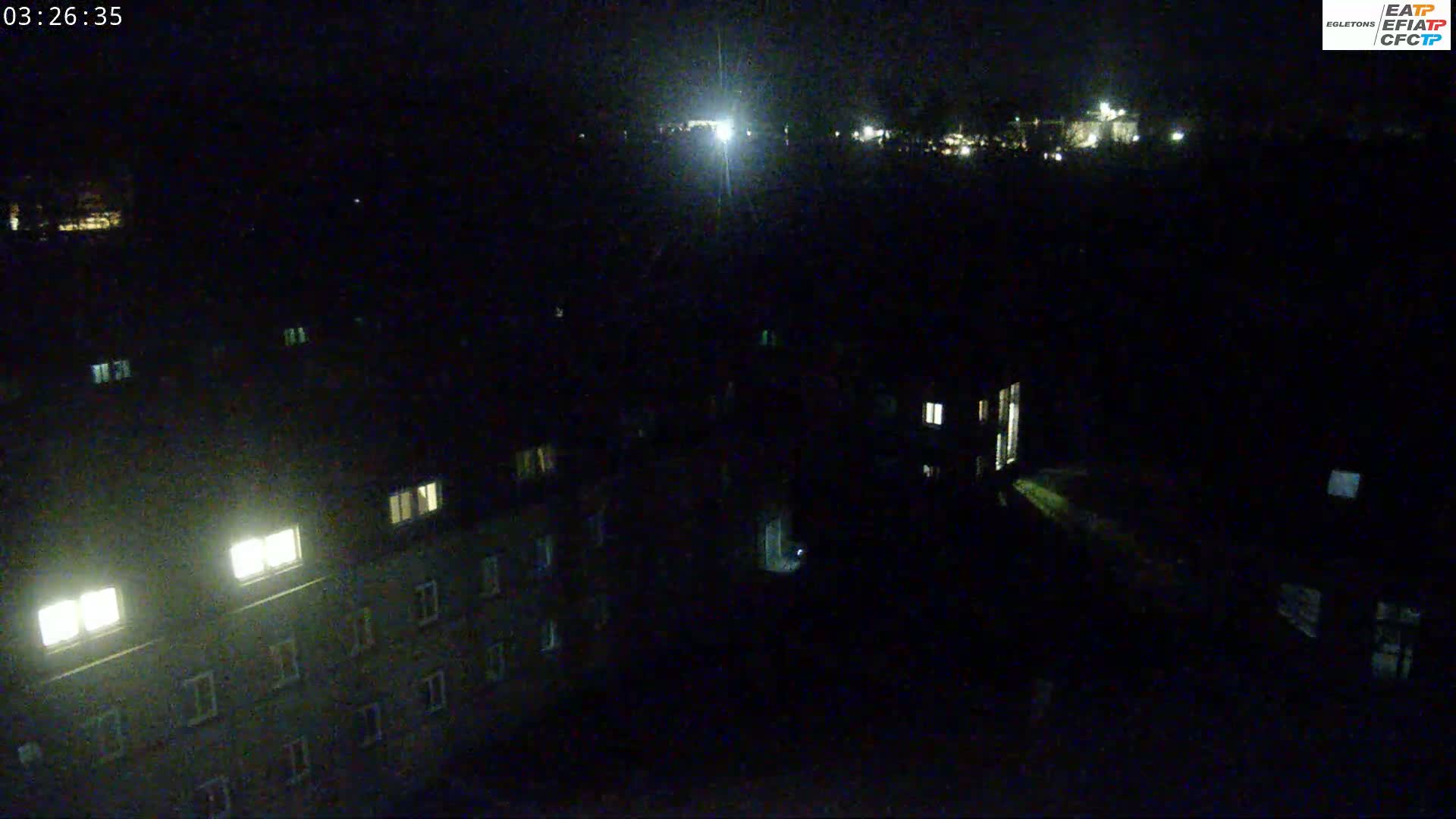 A nighttime, high-angle view shows a dark cityscape with a few illuminated windows in buildings, and a bright light source in the distance.