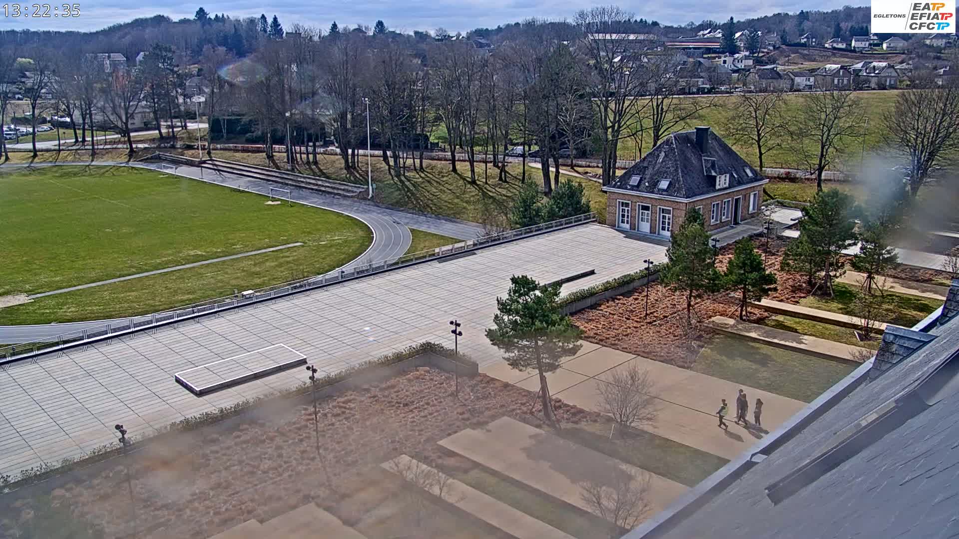 An aerial view shows a paved courtyard with a small building, a running track and soccer field, and a group of people walking in the courtyard on a sunny day.