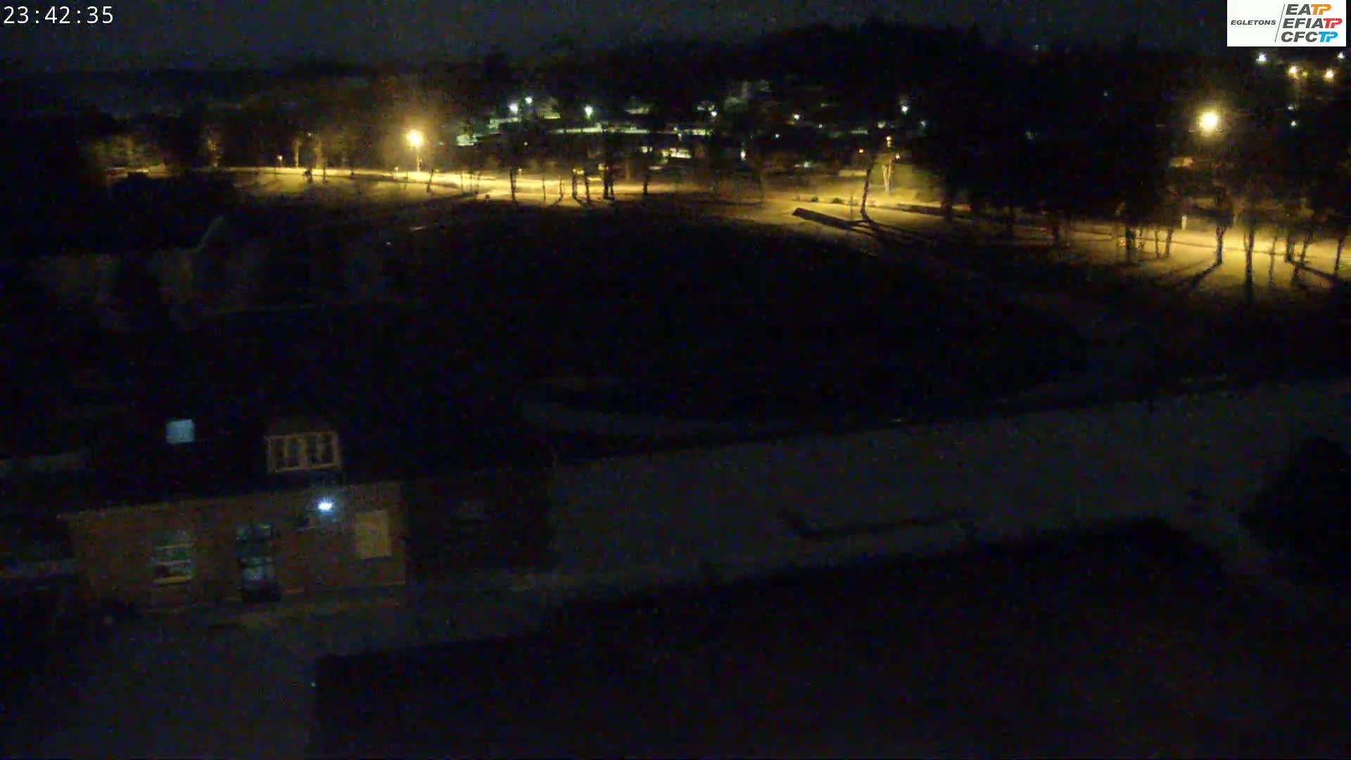 A nighttime view of a mostly dark, empty field, bordered by sparsely lit trees and buildings, under a clear night sky.