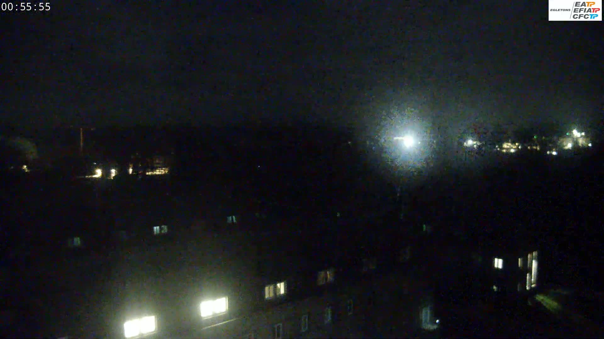 A nighttime view shows a dark landscape with a few lights from buildings and a bright light source in the distance, under a dark sky.