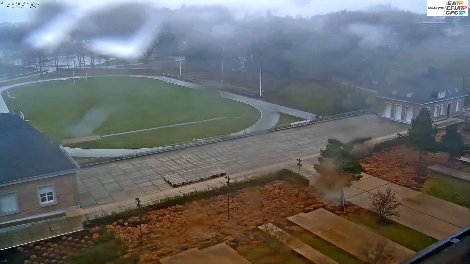 A high-angle view of a running track and field, a building, and landscaped grounds on a foggy and overcast day.
