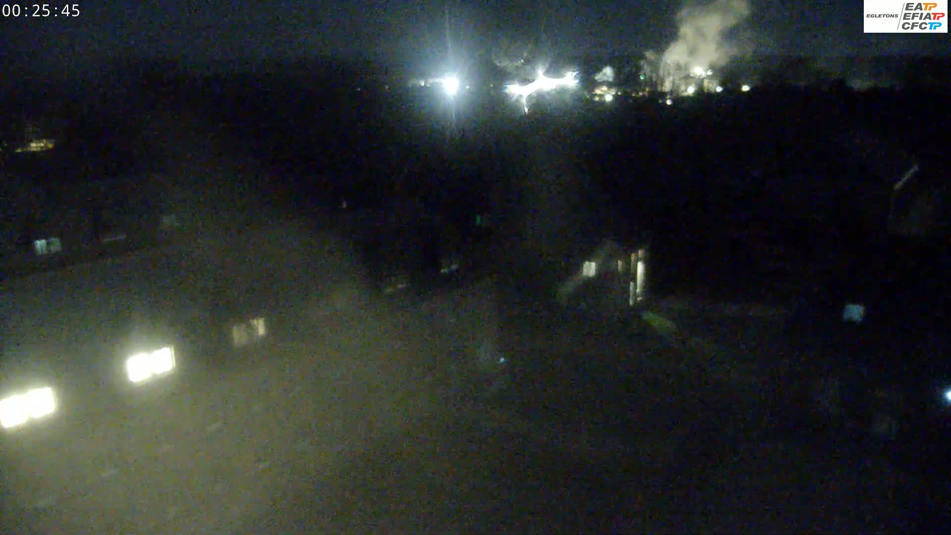 A nighttime, hazy view shows illuminated buildings and vehicles in the foreground with a distant industrial area emitting smoke under a dark sky.